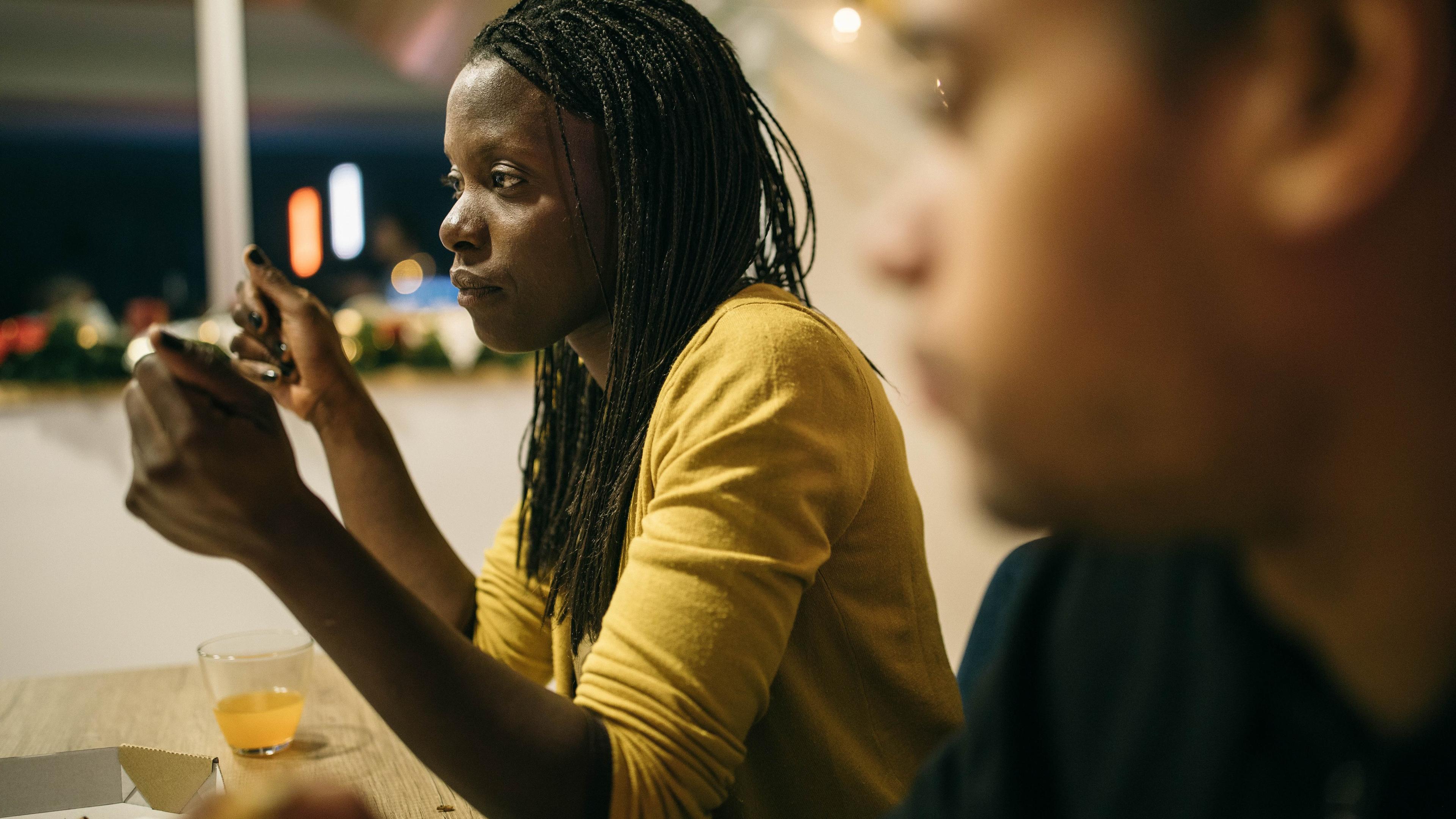 Photo of a woman in a yellow top using her phone at a table, with a blurred person in the foreground.