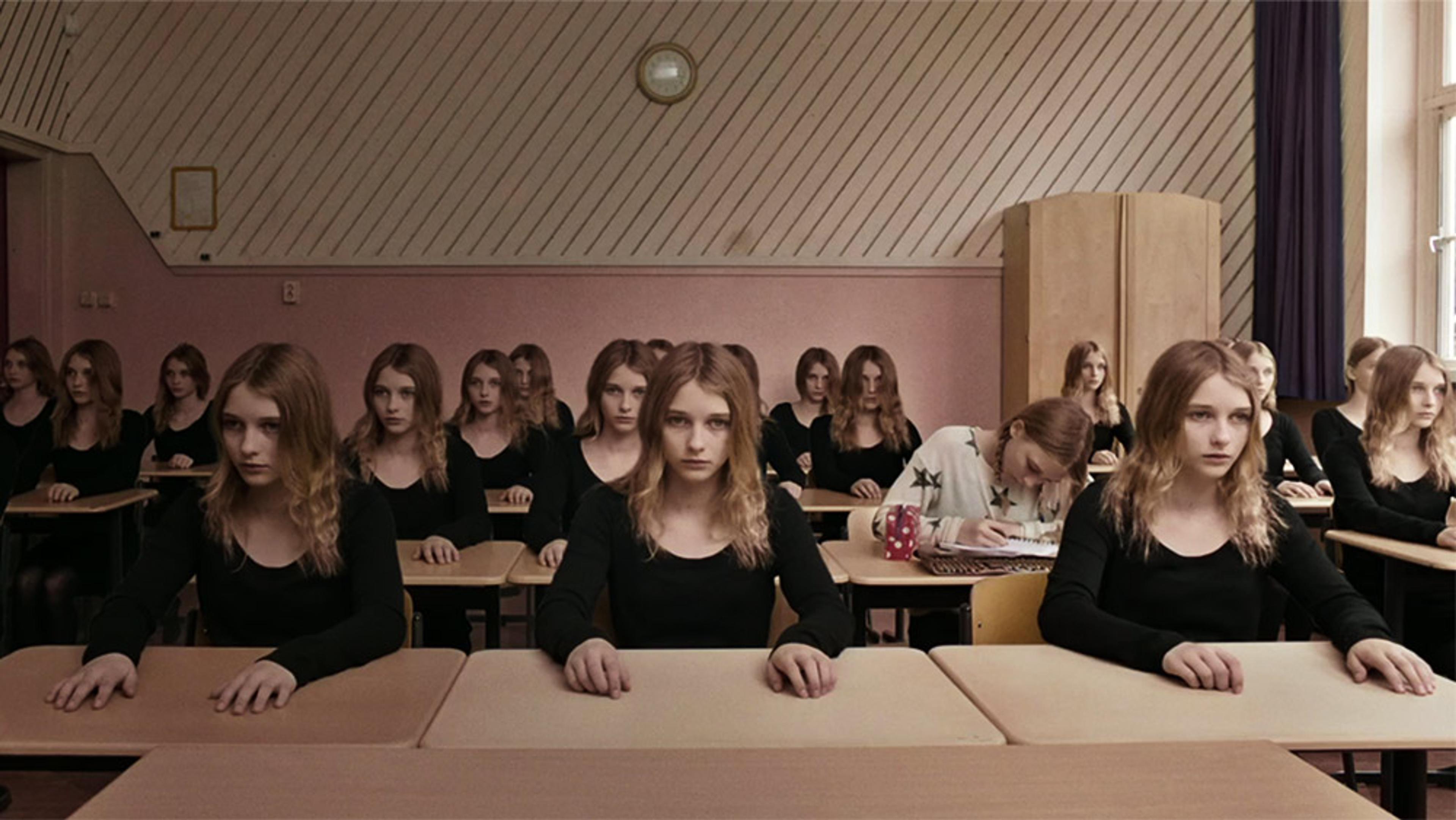 A classroom filled with identical girls in black tops, all looking ahead with sad eyes, with one girl in a white top decorated with stars looking down at her work.