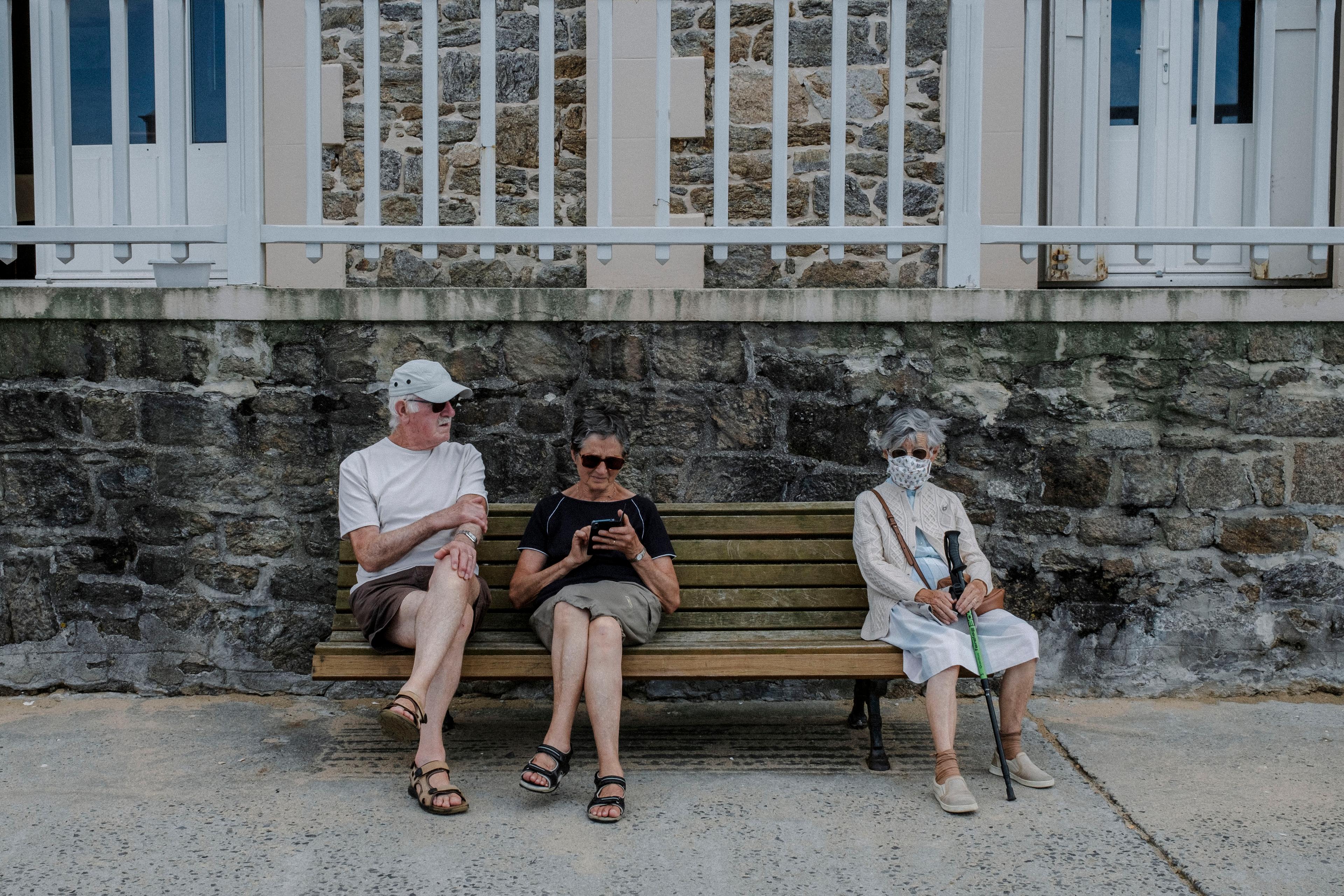 Photo of three elderly people sitting on a bench in front of a stone wall, one using a smartphone, another wearing a face mask.