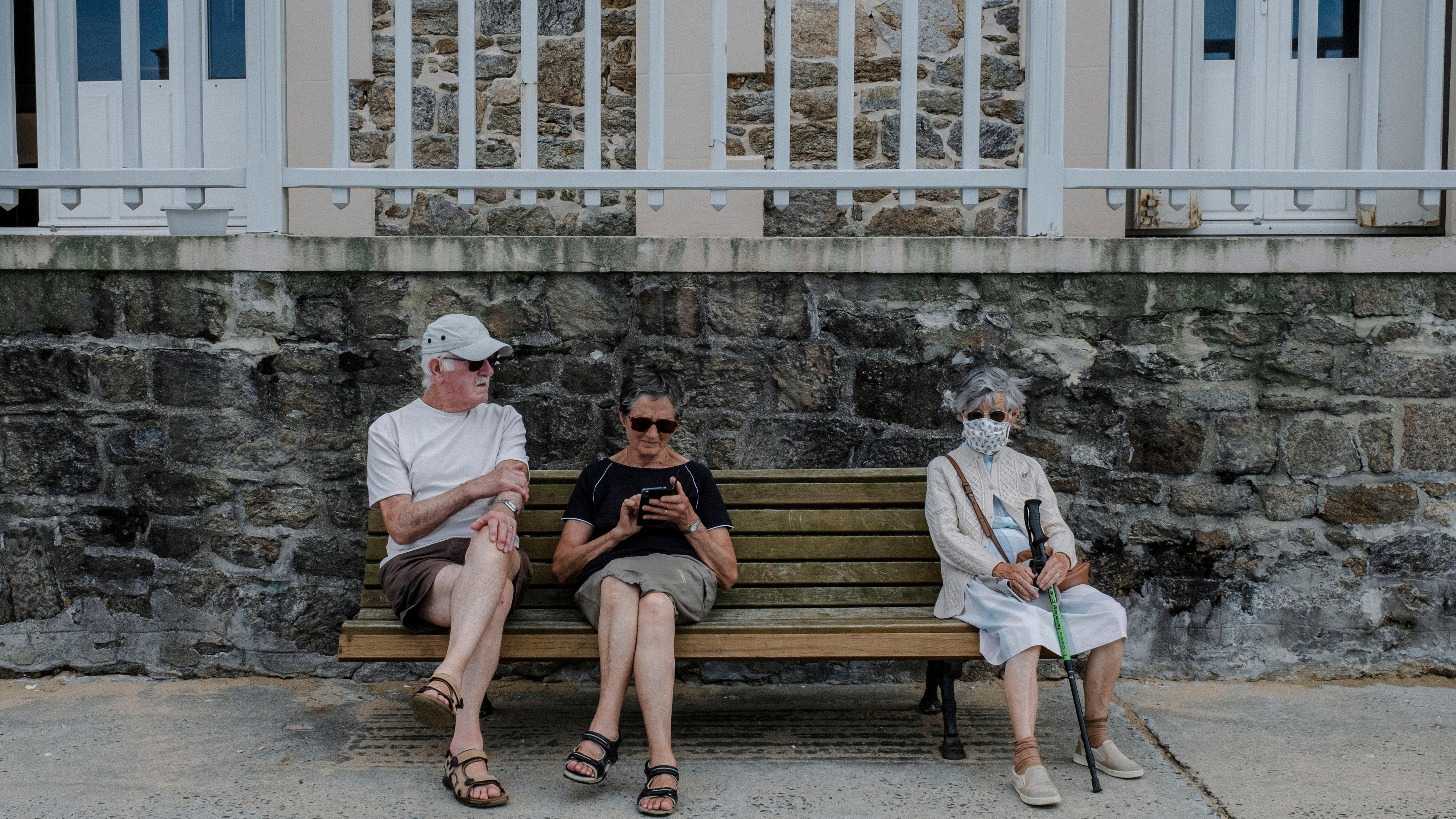 Photo of three elderly people sitting on a bench in front of a stone wall, one using a smartphone, another wearing a face mask.