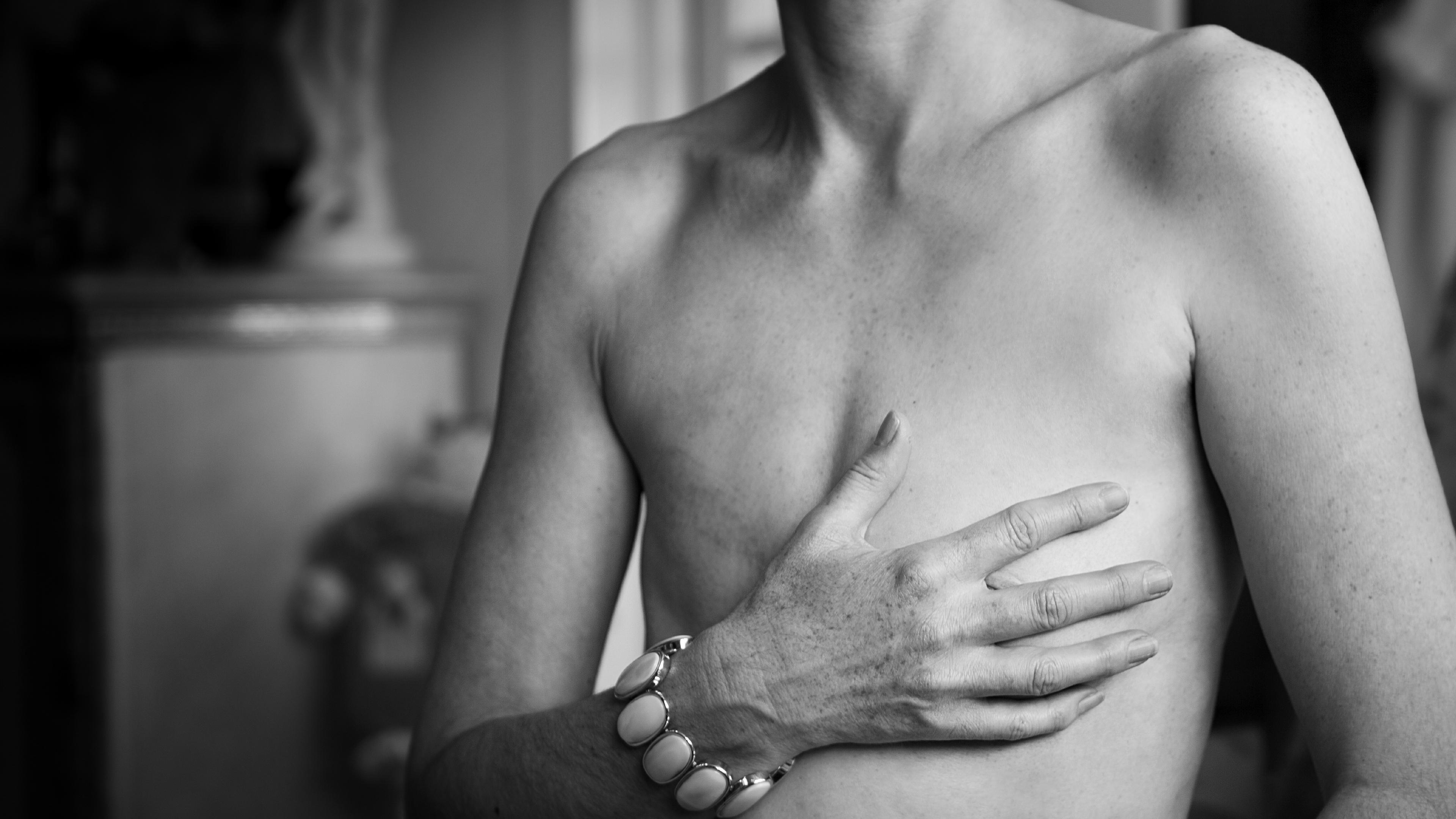 Black and white photo of a topless person covering their chest with a hand wearing a bracelet.