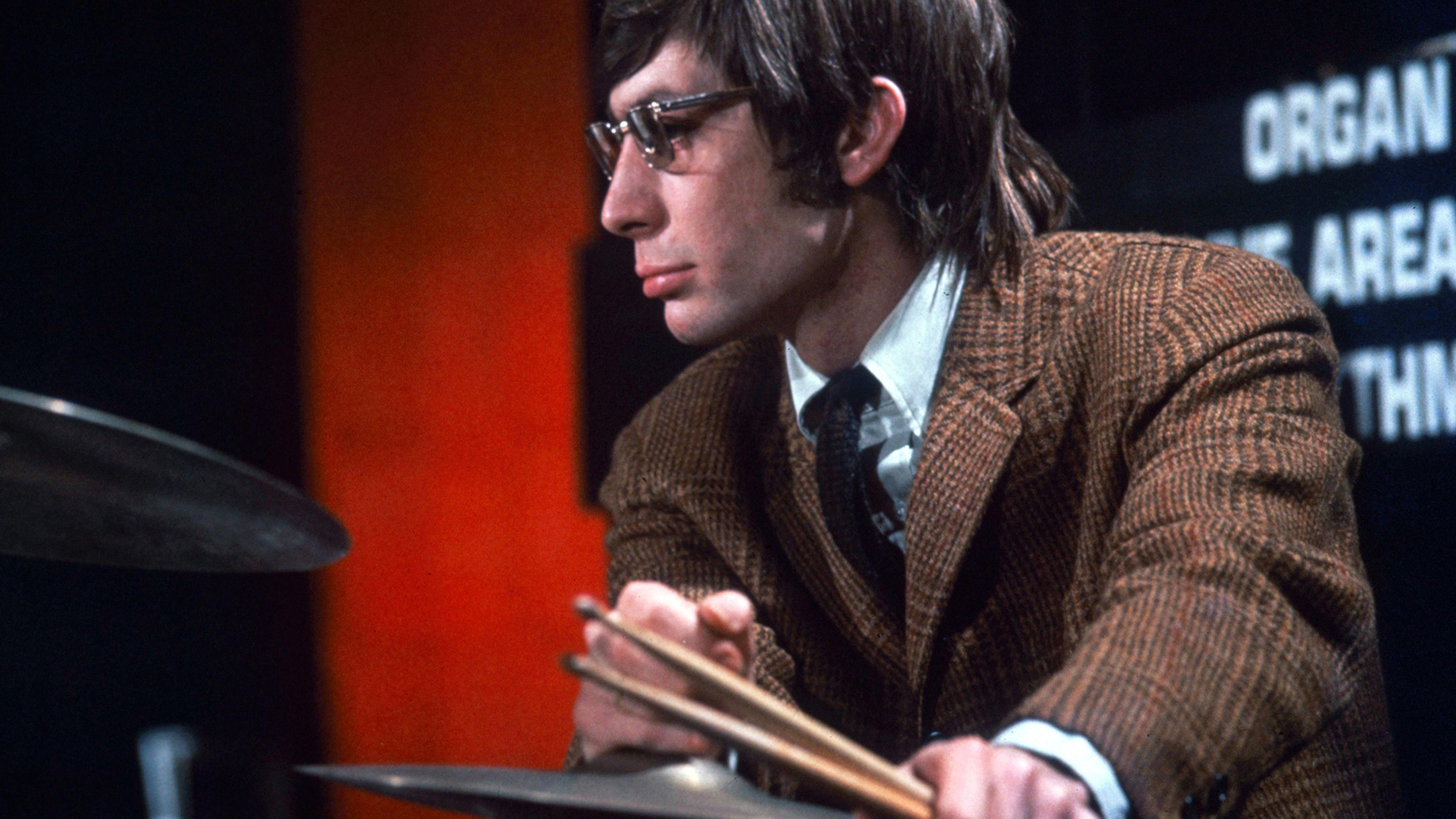 A man wearing glasses and a brown jacket who is Charlie Watts playing drums in a TV studio.