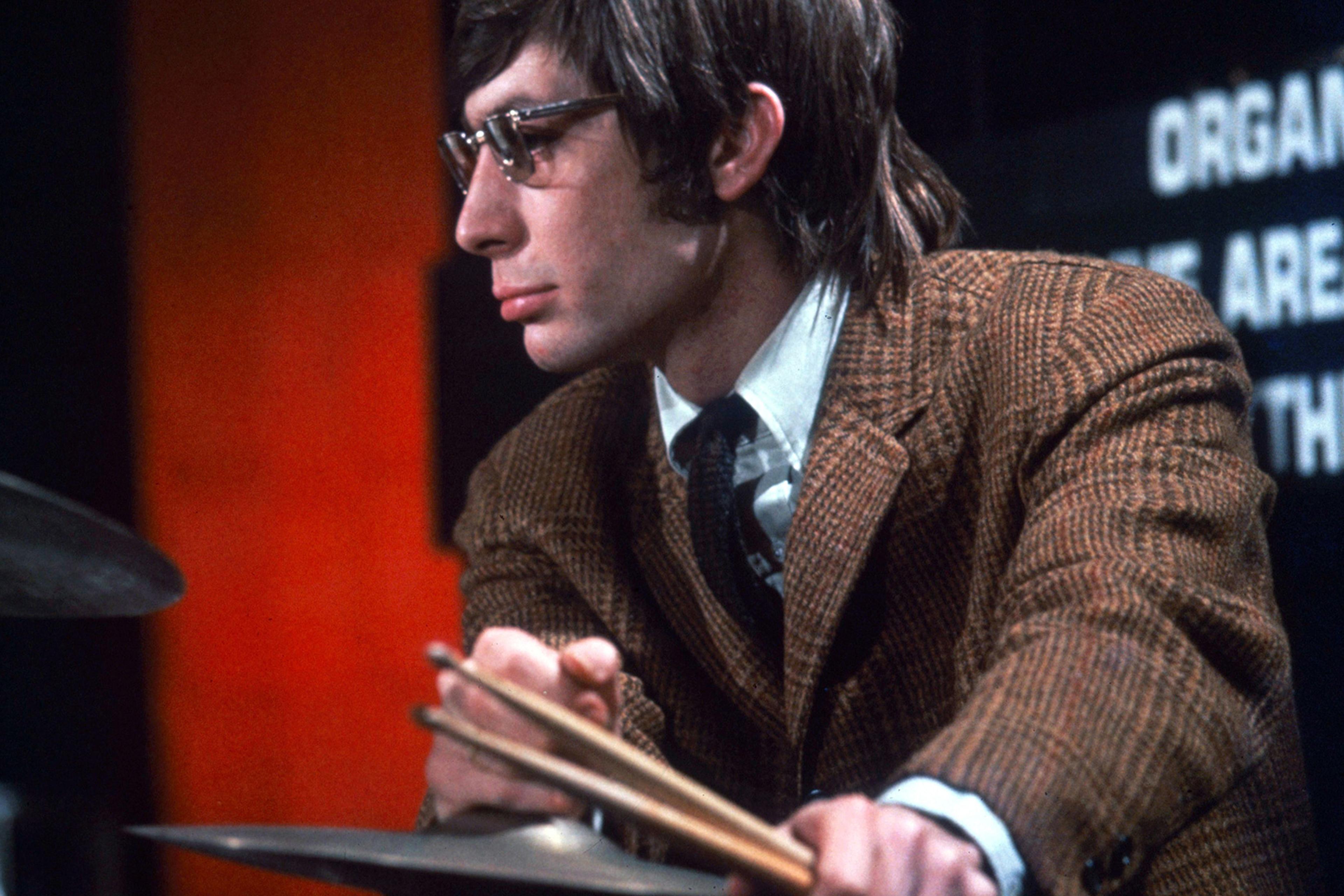 A man wearing glasses and a brown jacket who is Charlie Watts playing drums in a TV studio.