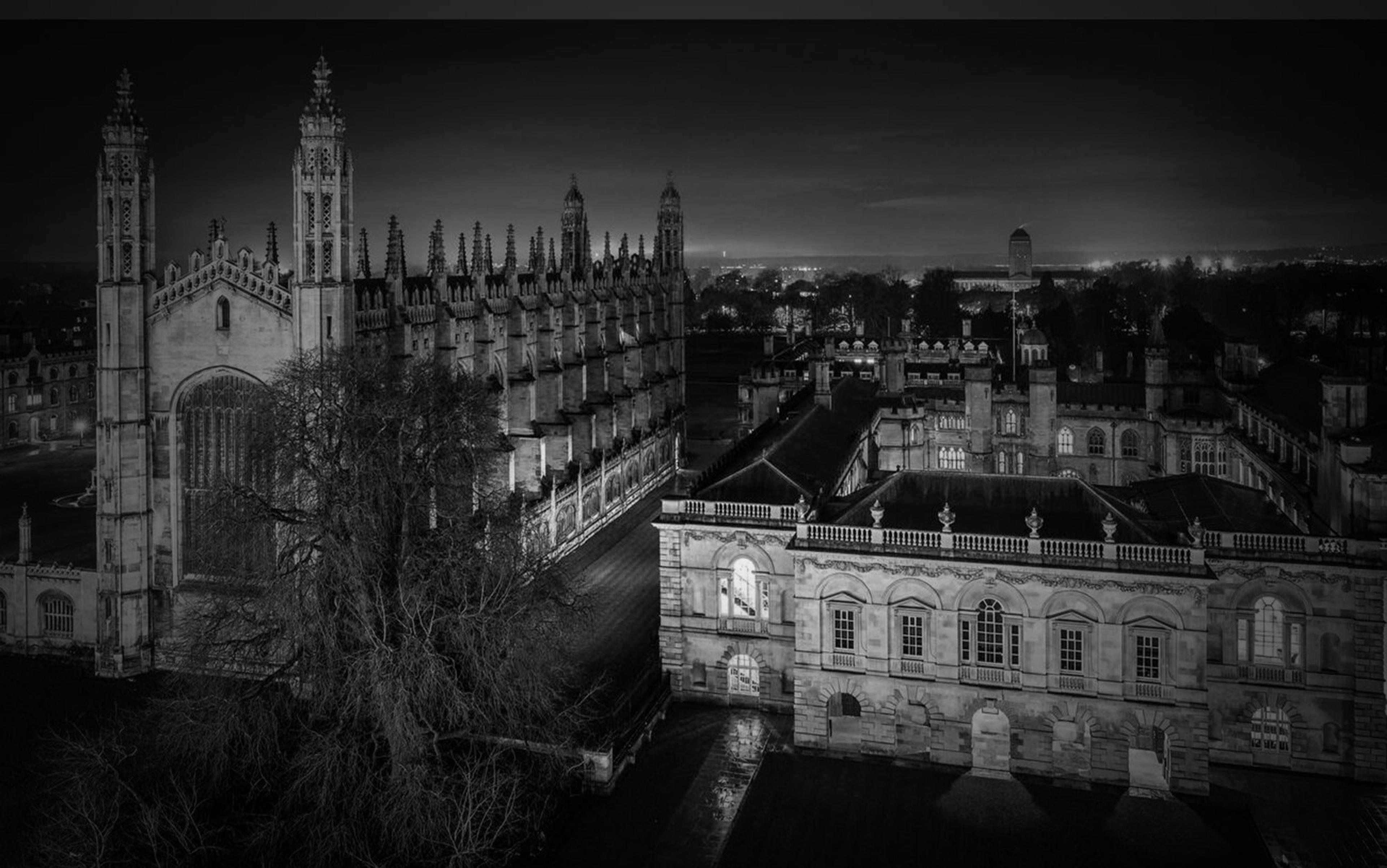 Black and white night photo of historic Gothic-style buildings with arched windows, towers and courtyards.