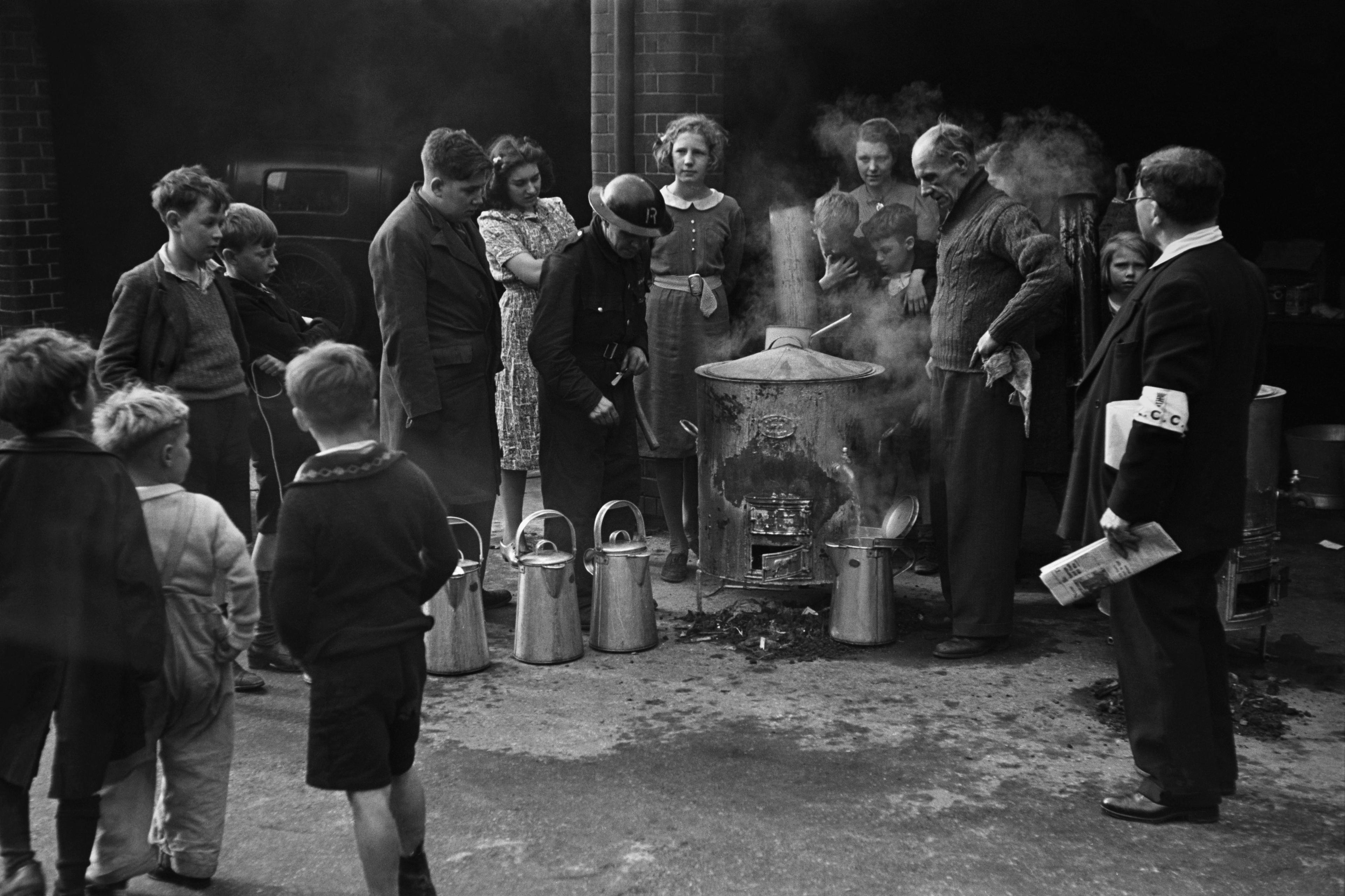 Black and white photo of people gathered around a large metal pot with steam in an outdoor setting.