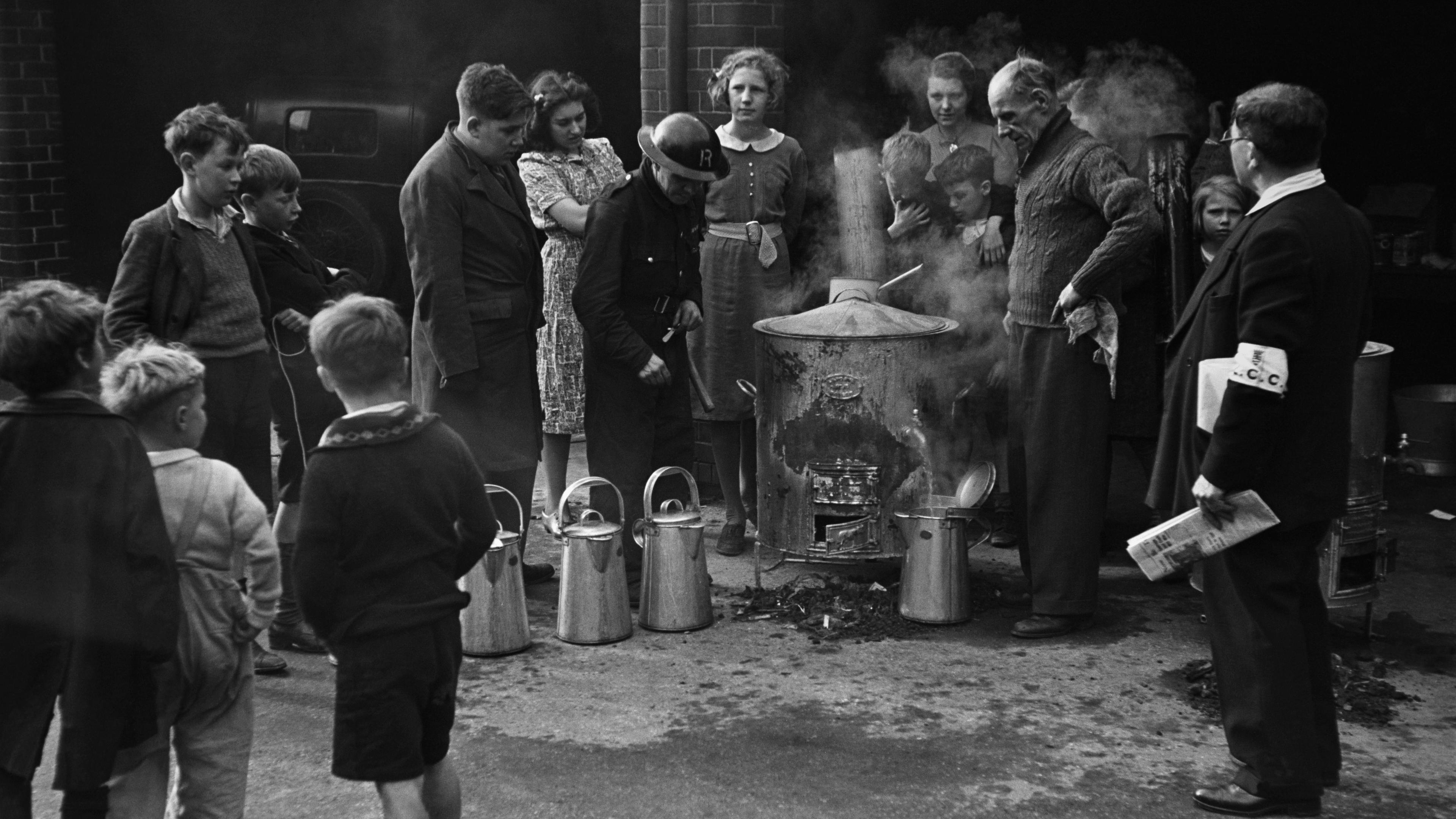 Black and white photo of people gathered around a large metal pot with steam in an outdoor setting.