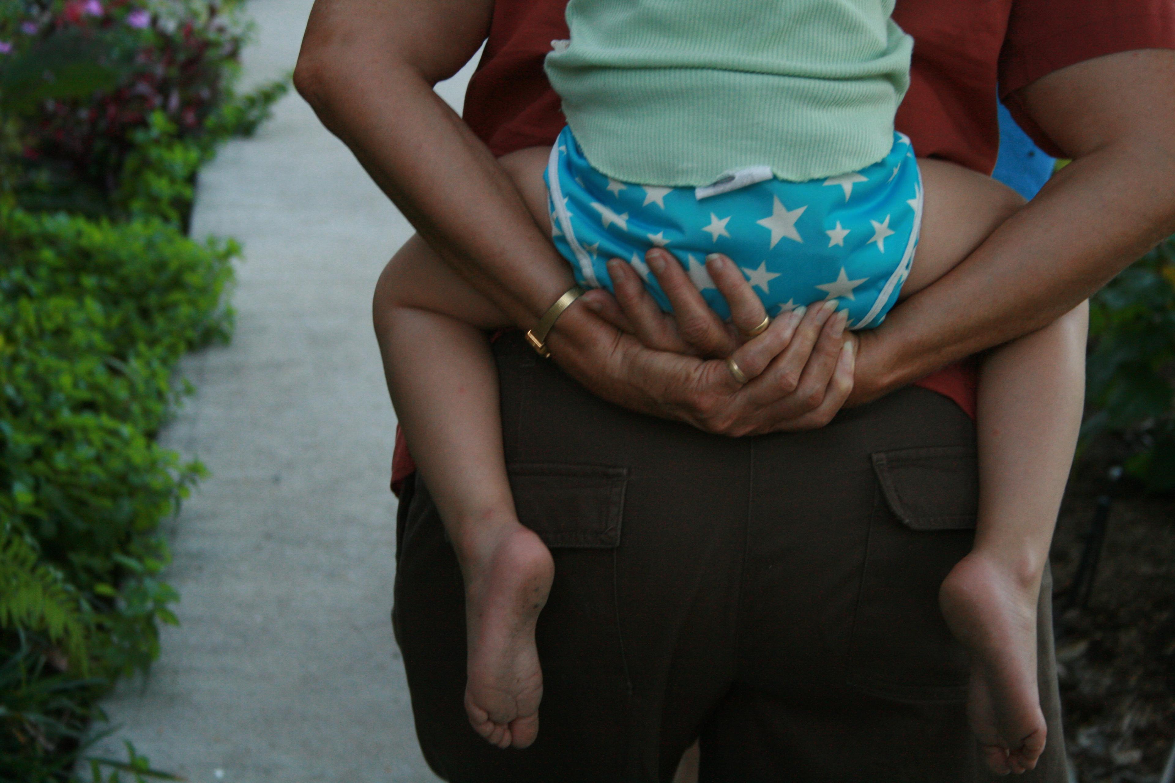 Photo of a person carrying a child with blue star shorts on their back, walking on a path lined with green plants.
