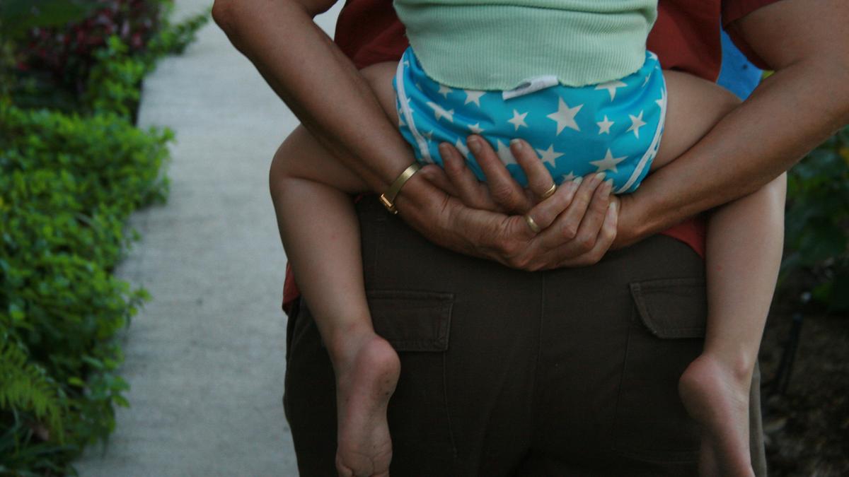 Photo of a person carrying a child with blue star shorts on their back, walking on a path lined with green plants.