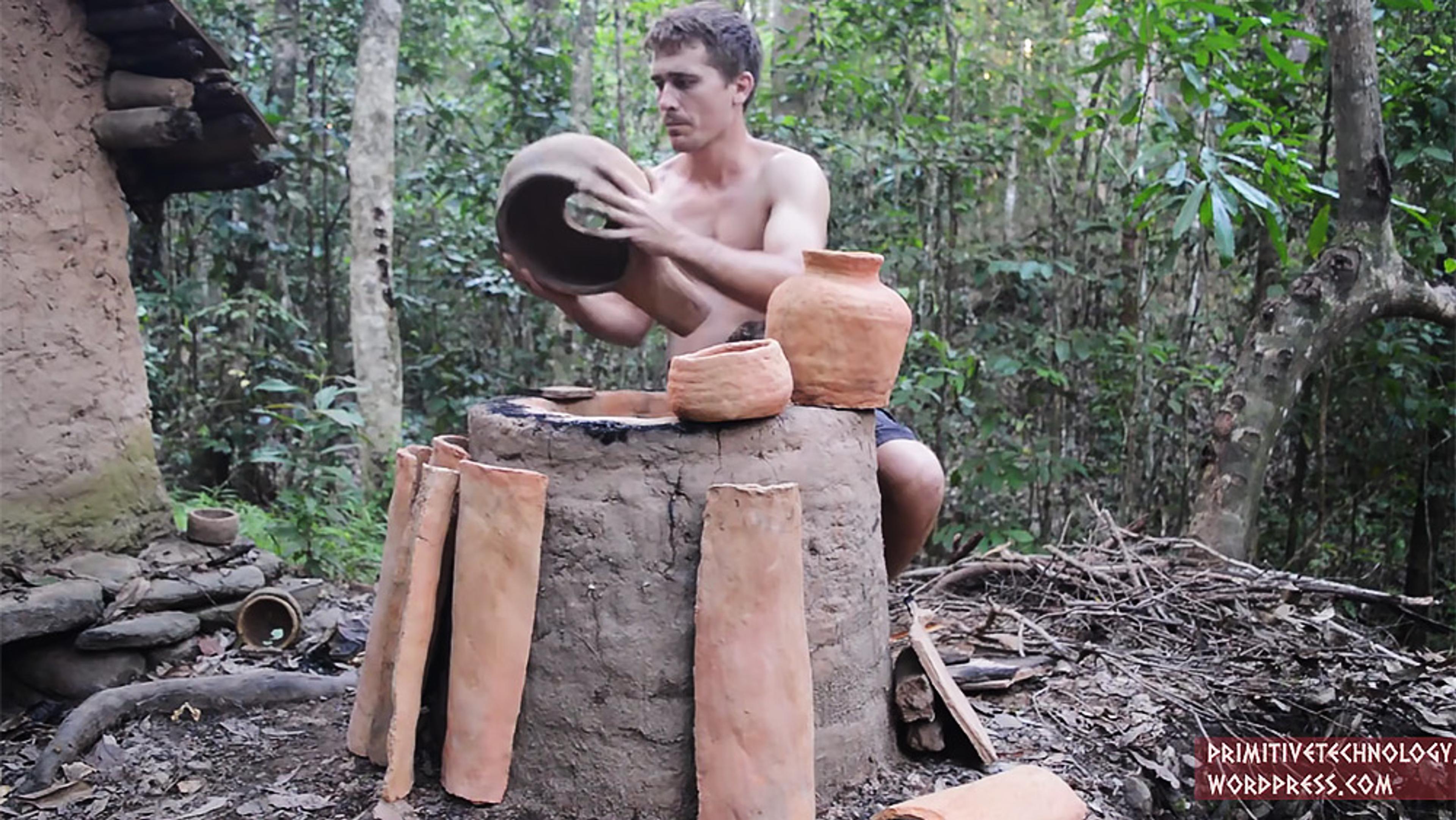 A shirtless man works on clay pottery beside a mud hut in a forest. Several pots and clay items are around him.