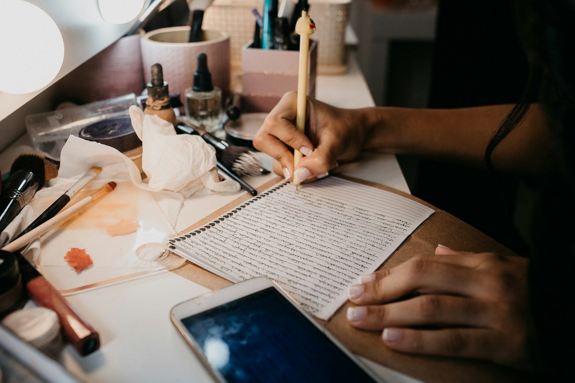 A person writing in a notebook on a cluttered vanity with makeup items and a smartphone nearby.