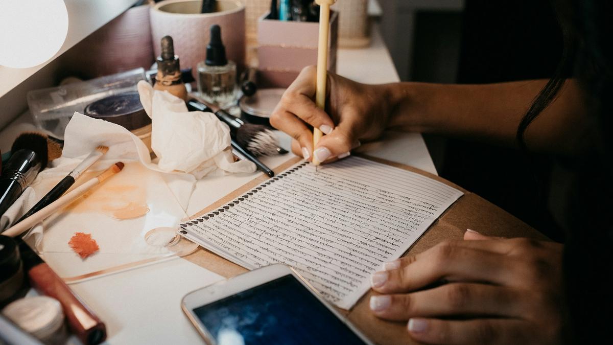 A person writing in a notebook on a cluttered vanity with makeup items and a smartphone nearby.
