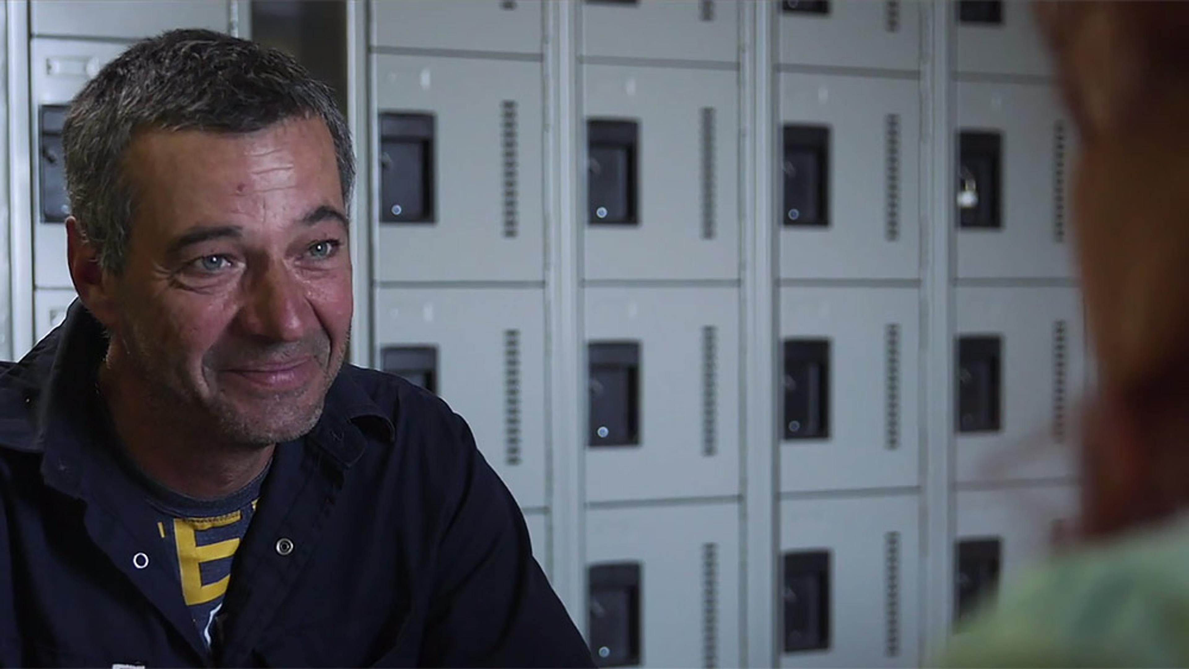 A man with grey hair and a slight smile. He is in a room with rows of grey lockers, speaking to an unseen person.