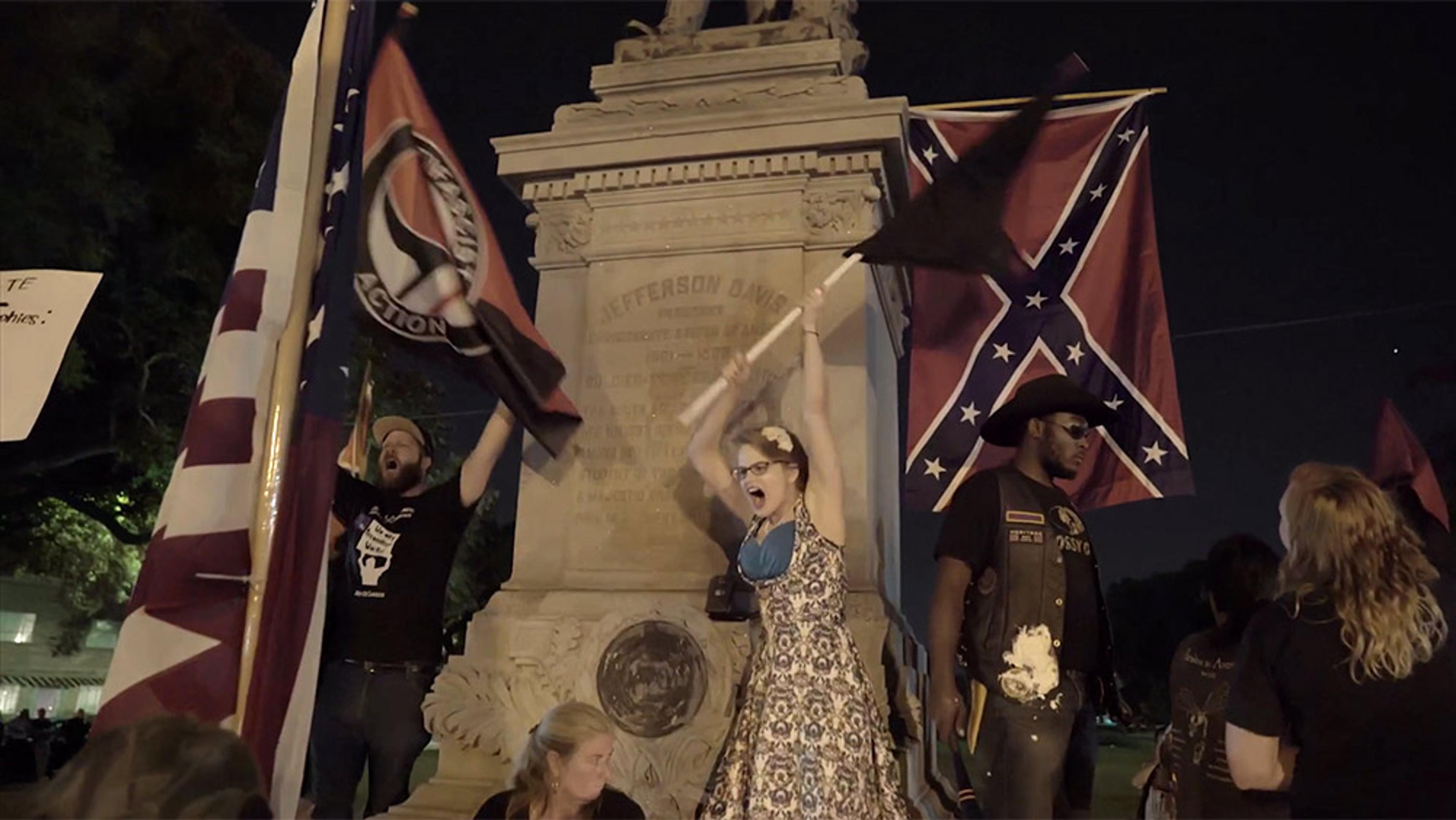 Multiple people protesting at a Jefferson Davis monument, holding various flags including an American flag and a Confederate flag.