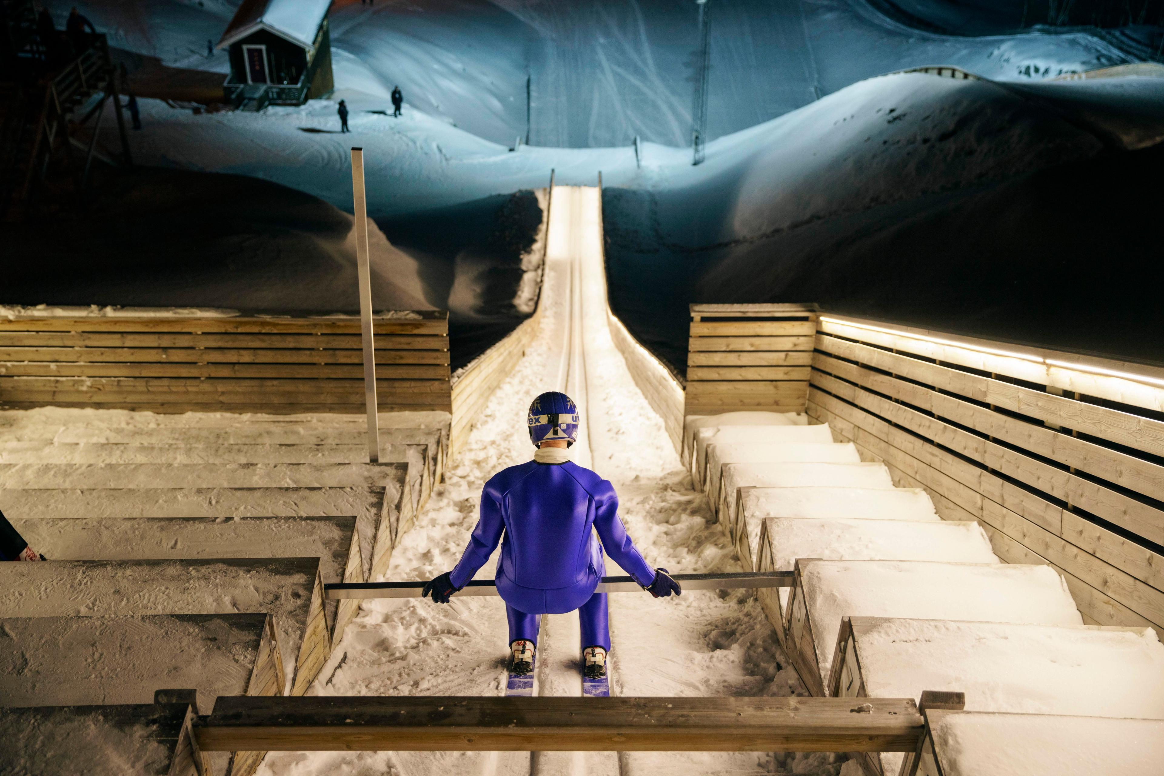 Photo of a ski jumper in blue gear at the top of a snow-covered ramp preparing to jump at night.
