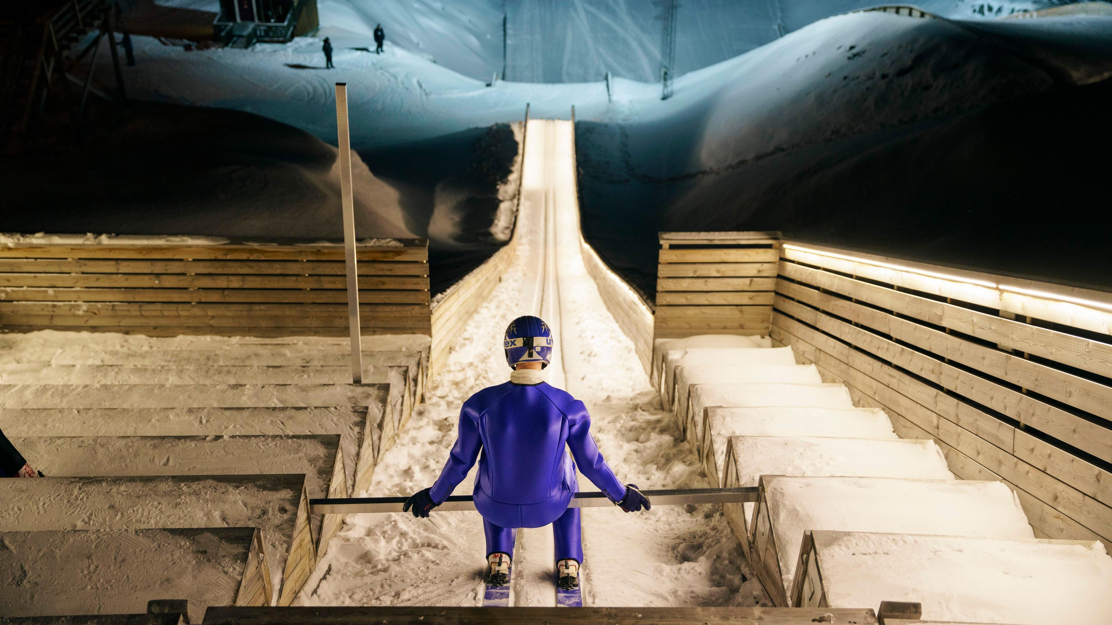 Photo of a ski jumper in blue gear at the top of a snow-covered ramp preparing to jump at night.