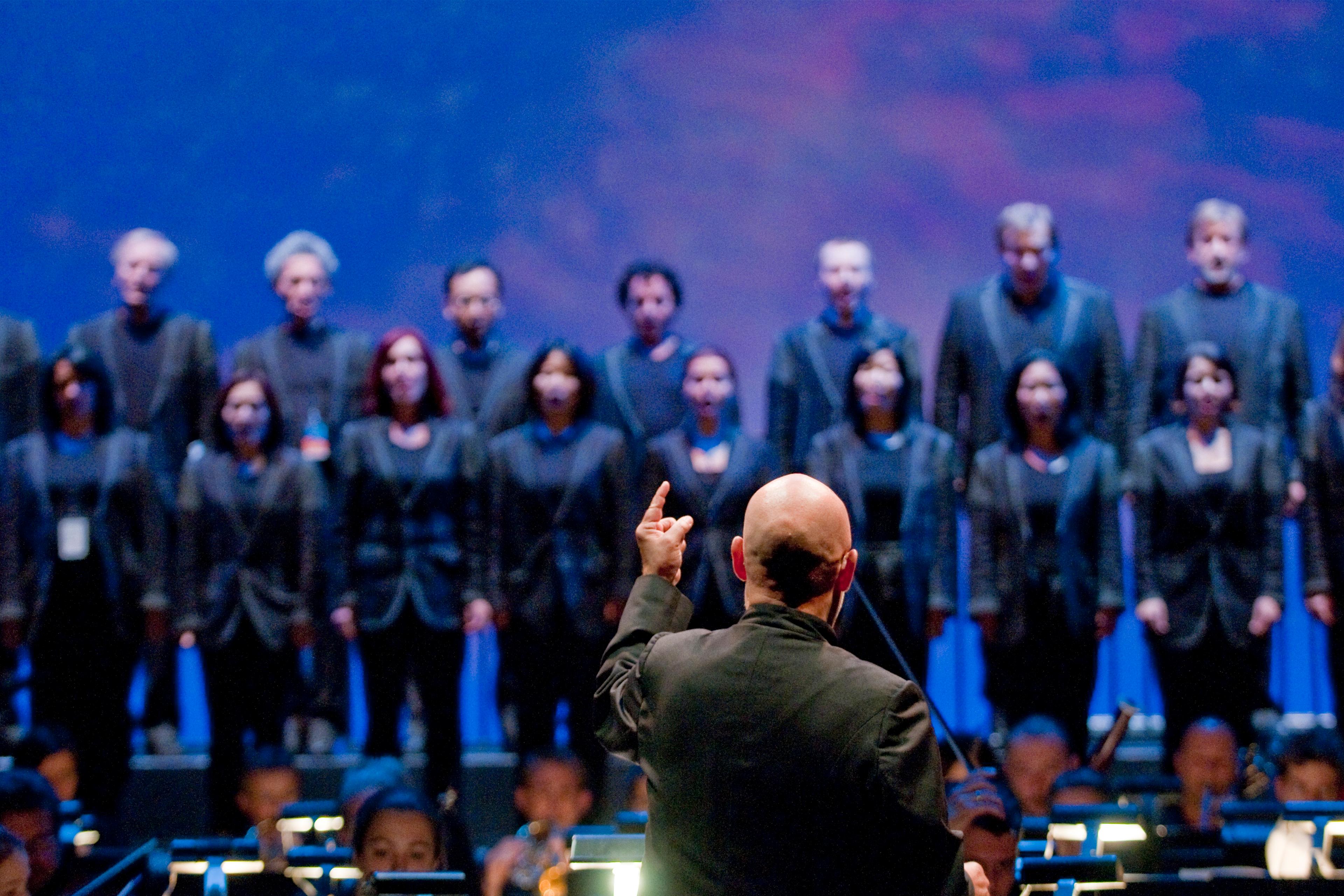 Photo of a conductor leading a choir on stage, blue background, choir members in black attire, focus on conductor’s back.