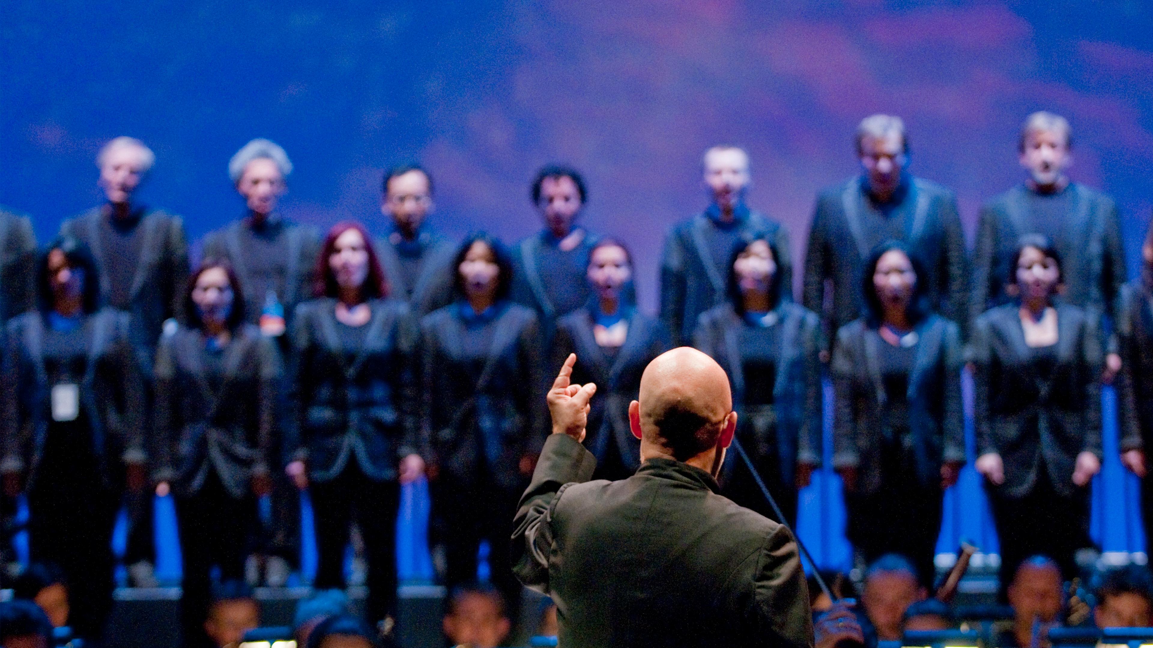 Photo of a conductor leading a choir on stage, blue background, choir members in black attire, focus on conductor’s back.