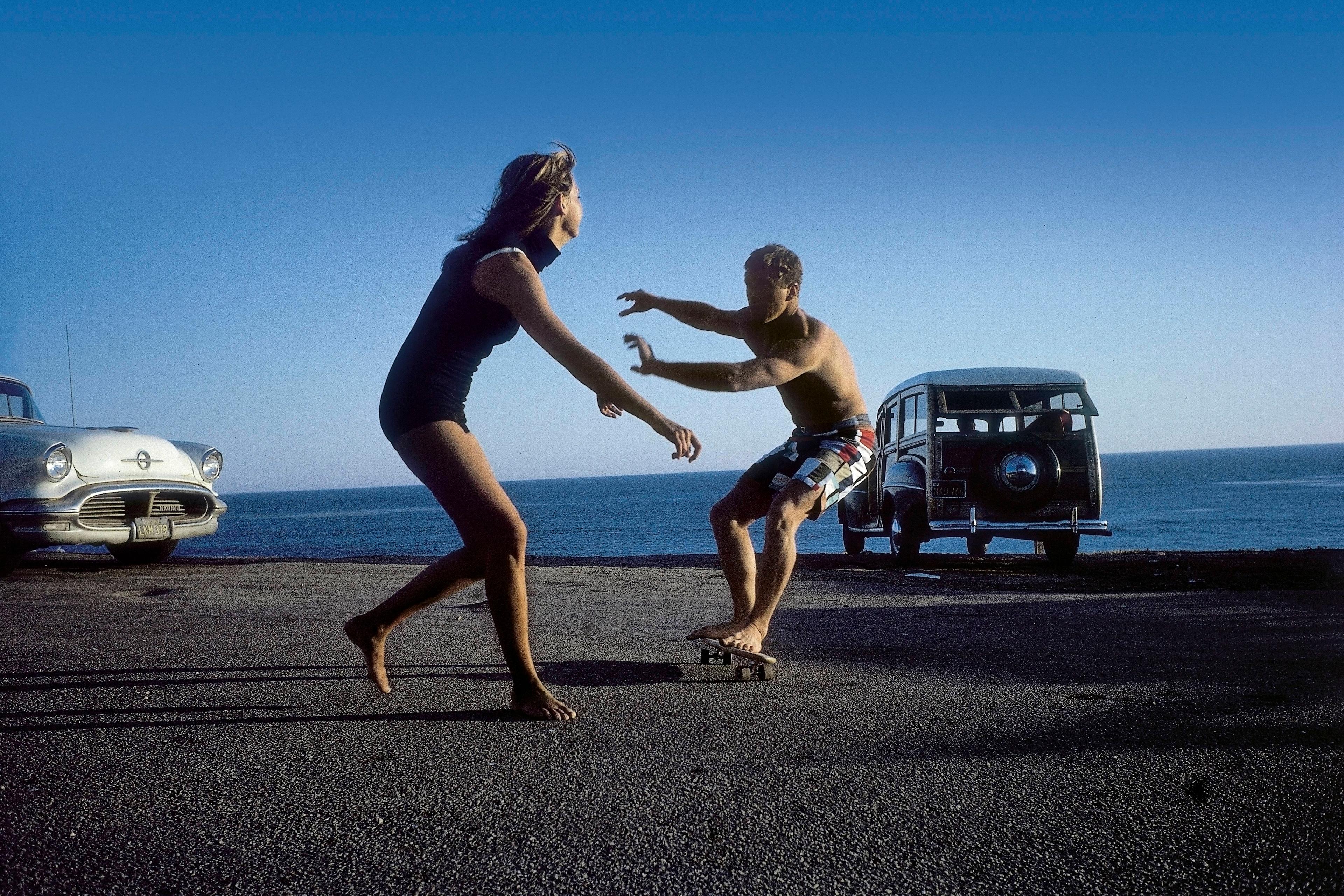 Photo of a man skateboarding beside the ocean with an old car watching a woman in a swimsuit on a sunny day.