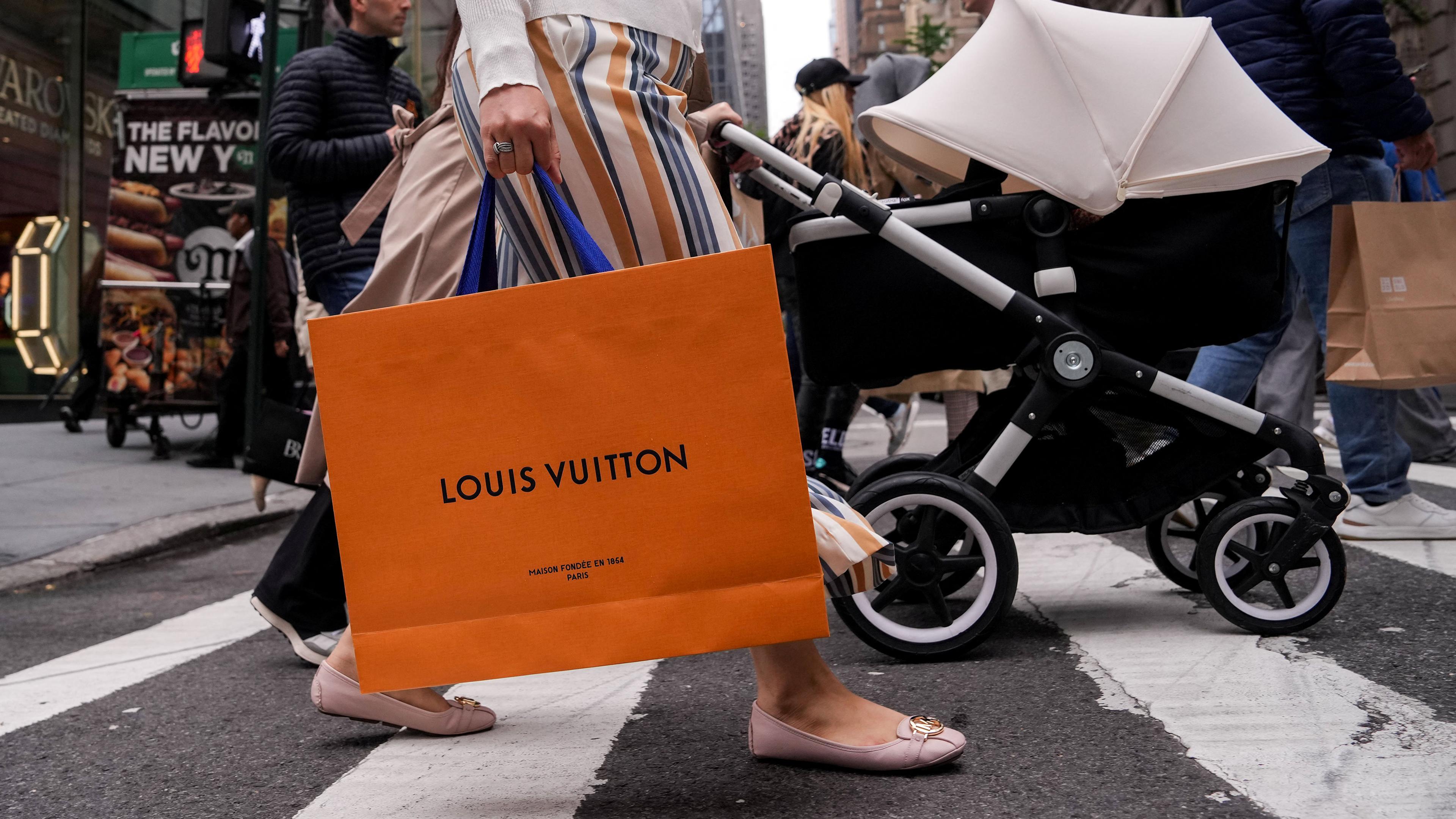 A person crossing a busy city street holding a Louis Vuitton bag with other pedestrians in the background.