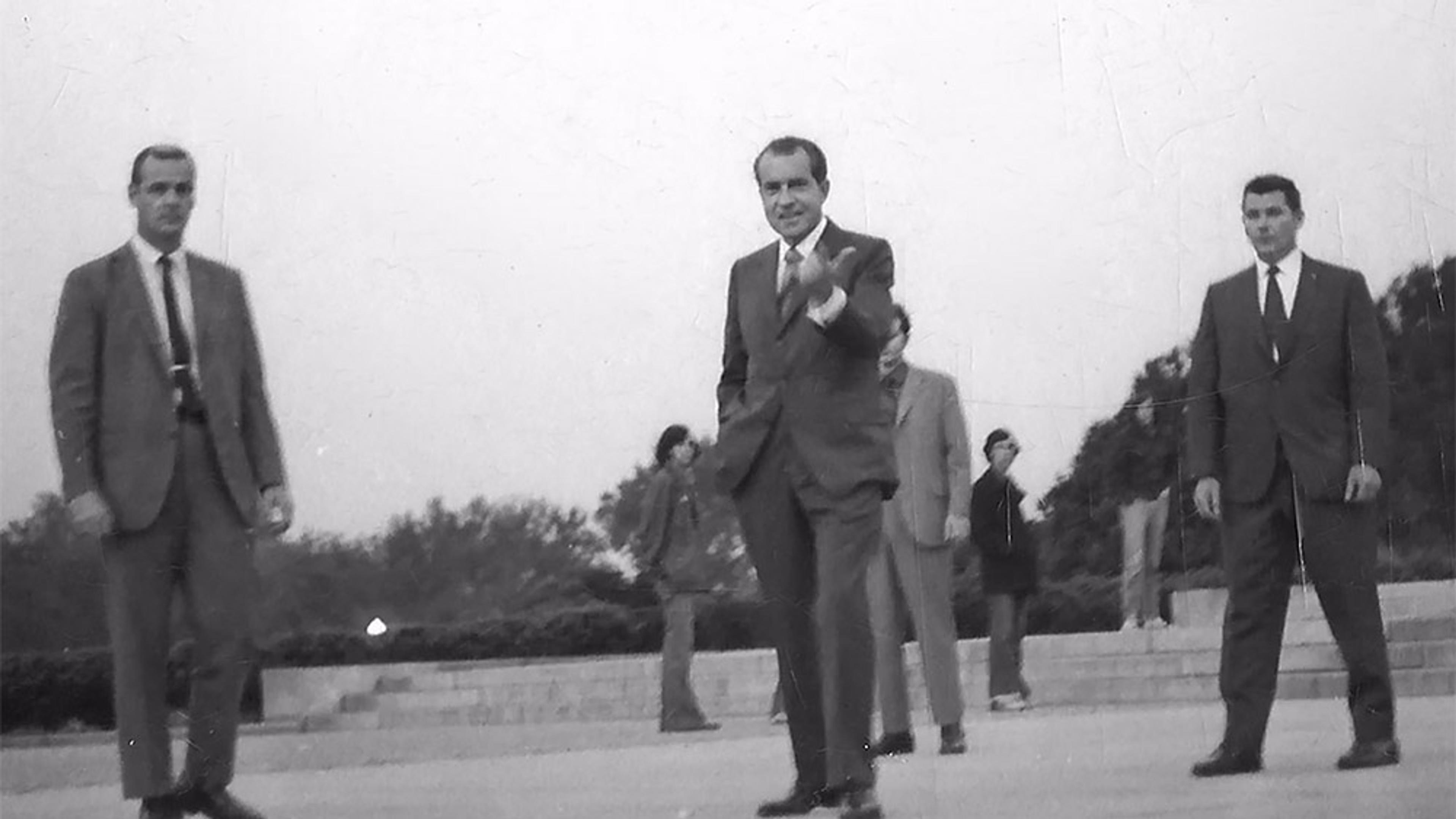 Black and white photo of a group of men in suits outdoors, the man in the centre is gesturing, trees and steps in the background.