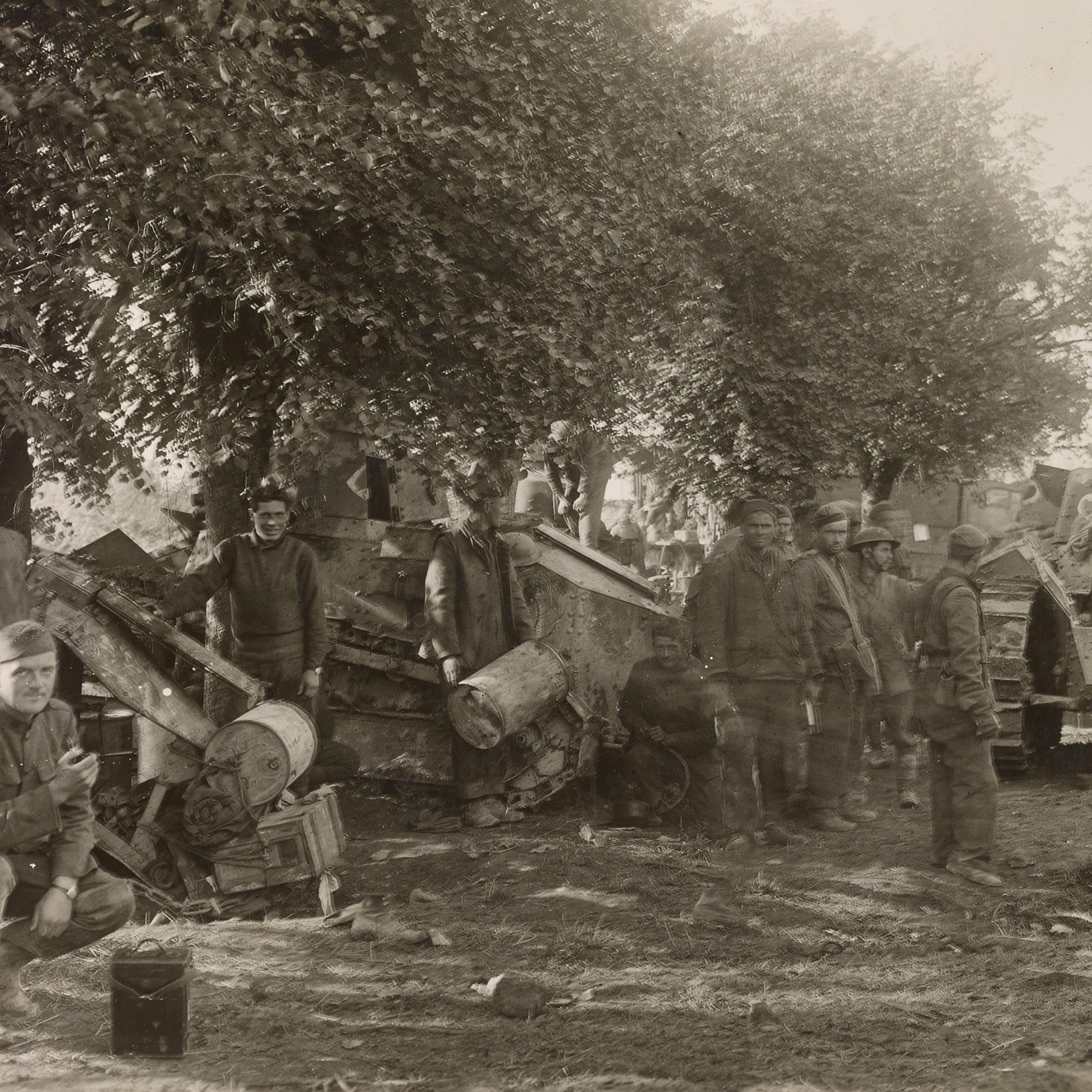 Black and white photo of soldiers and damaged tanks under trees in a wartime setting. One soldier crouches in the foreground.