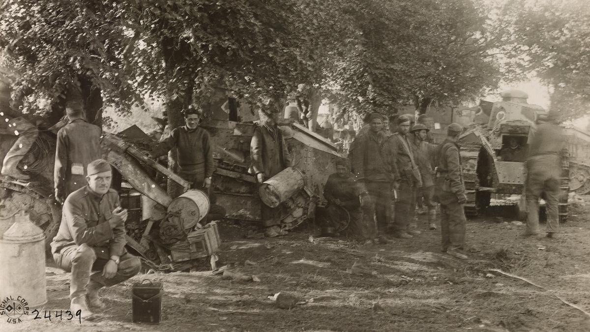 Black and white photo of soldiers and damaged tanks under trees in a wartime setting. One soldier crouches in the foreground.