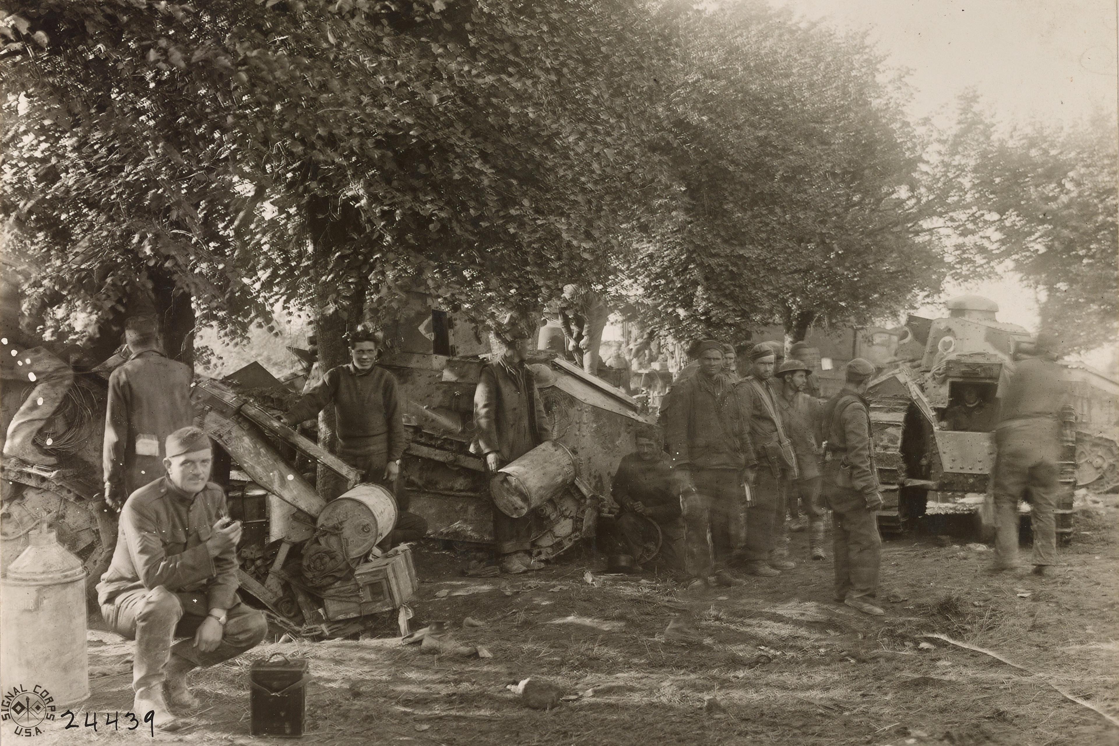 Black and white photo of soldiers and damaged tanks under trees in a wartime setting. One soldier crouches in the foreground.
