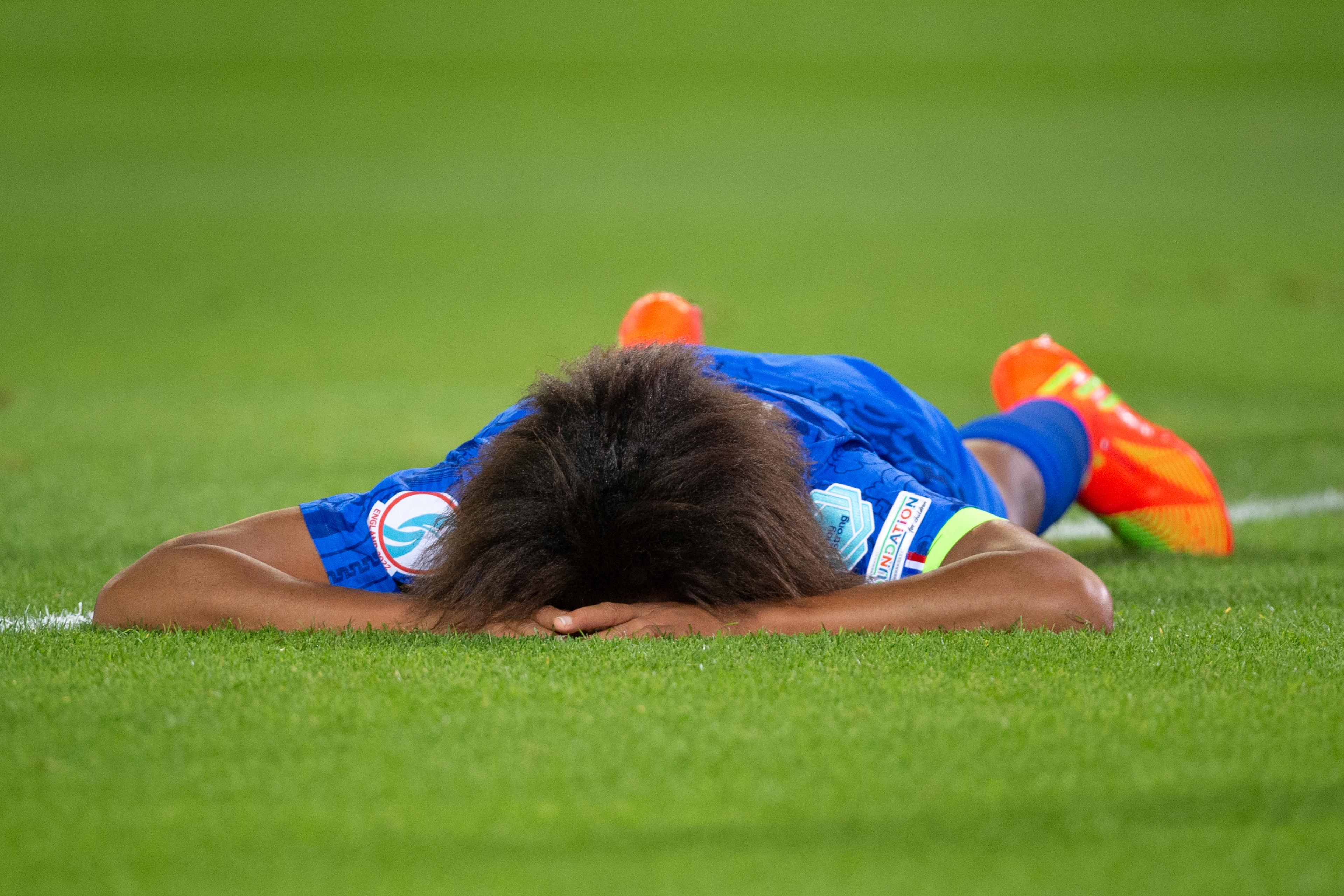 Photo of a footballer lying face down on the pitch in a blue kit with bright orange boots, hands covering their face.