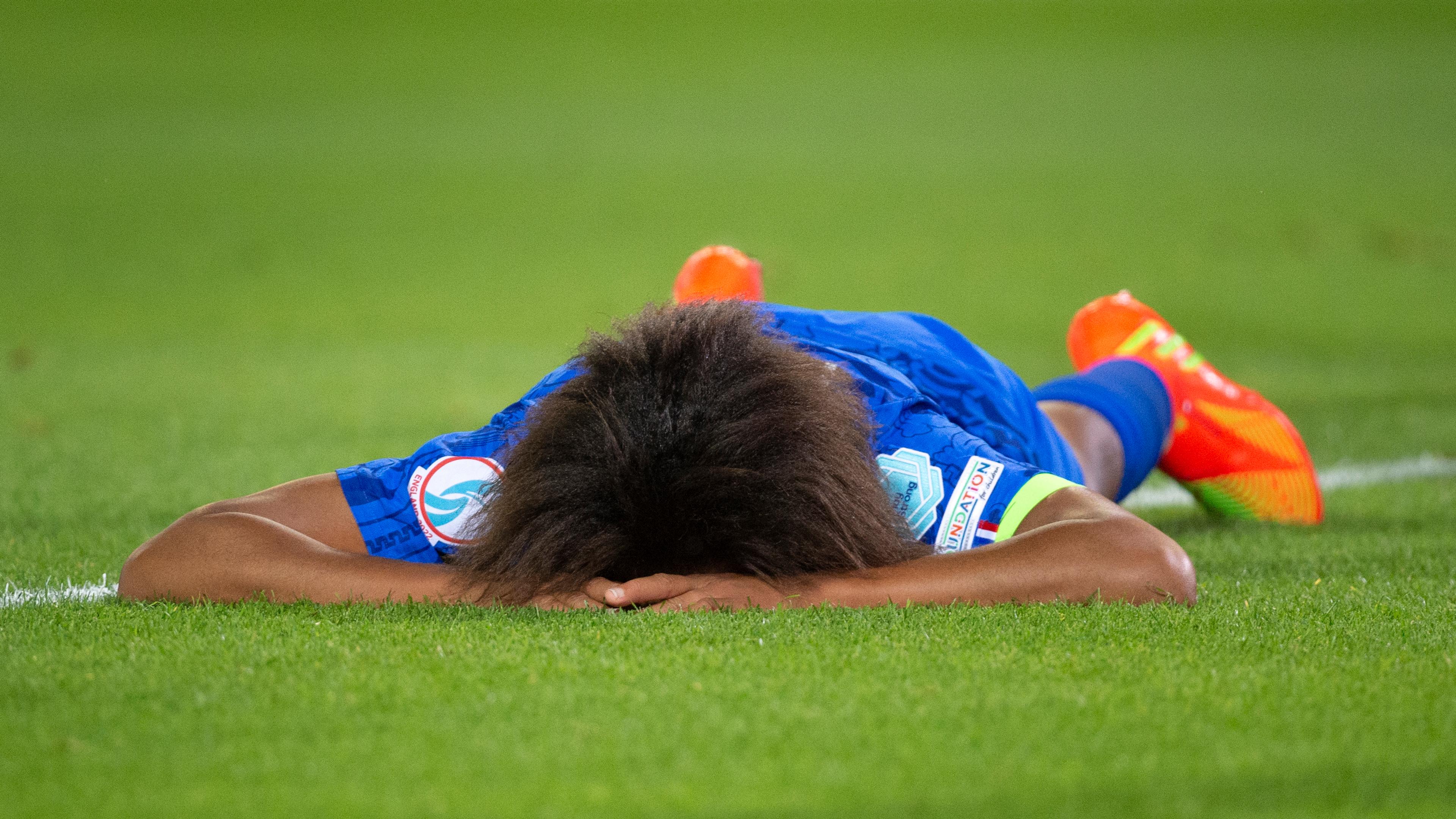 Photo of a footballer lying face down on the pitch in a blue kit with bright orange boots, hands covering their face.