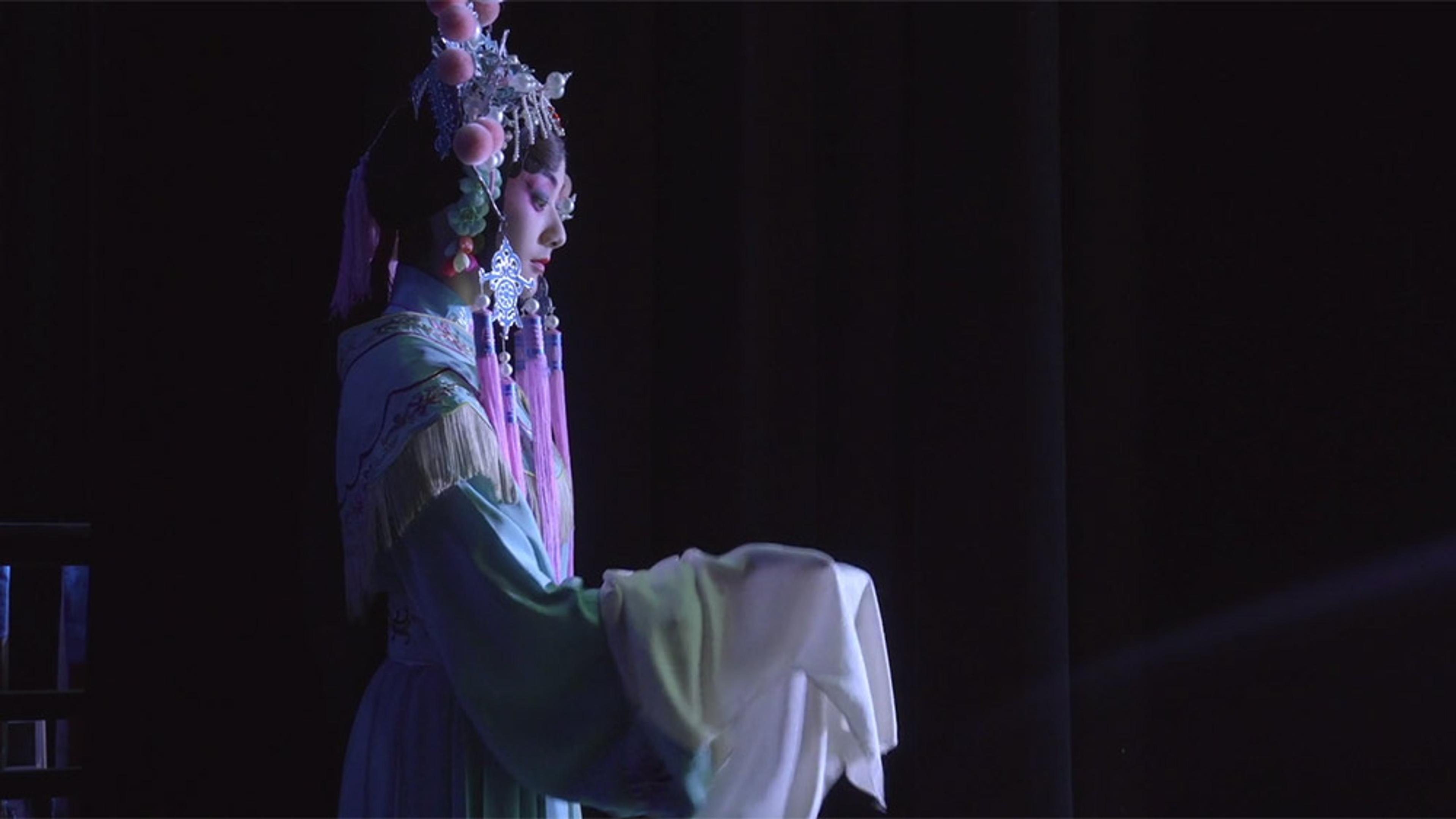 A performer in traditional Chinese opera attire and headpiece, standing in profile on a dimly lit stage holding white sleeves.