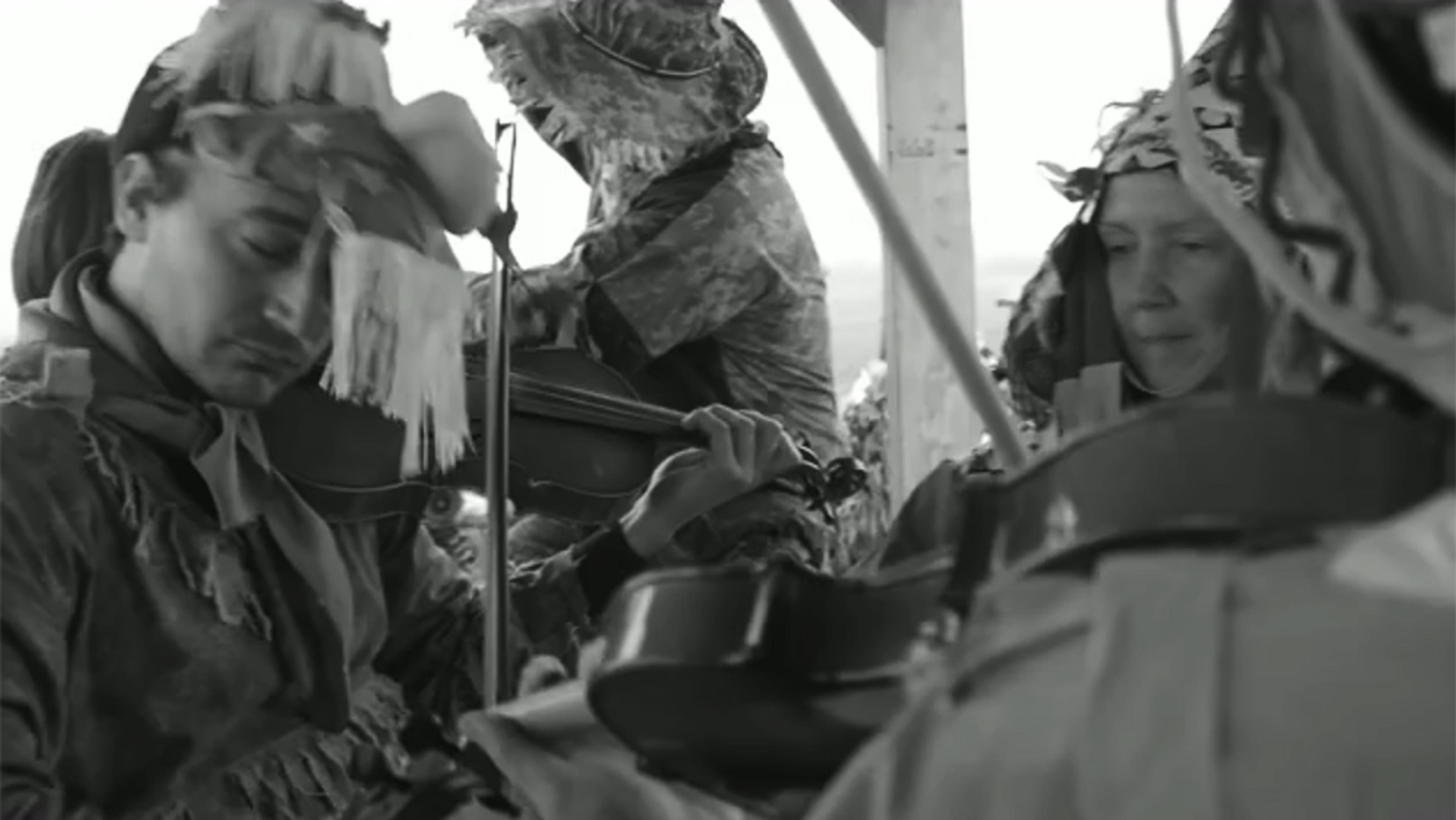Black and white photo of four people in costumes playing violins. The focus is on two individuals who are deeply engaged in their music.