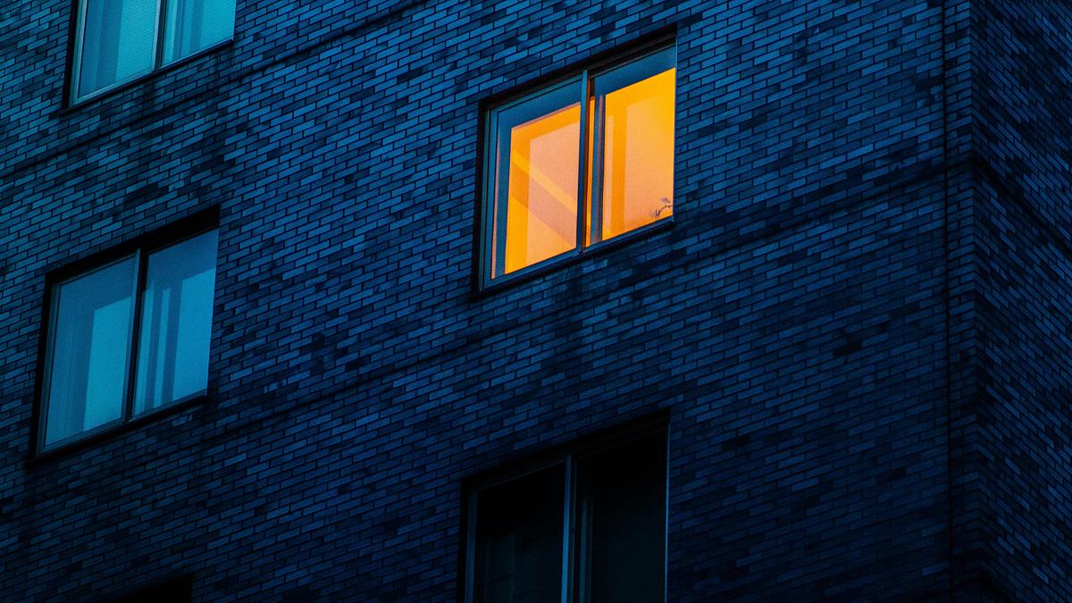 A dark building with a brightly lit orange window at night, surrounded by dimmer blue-tinted windows.