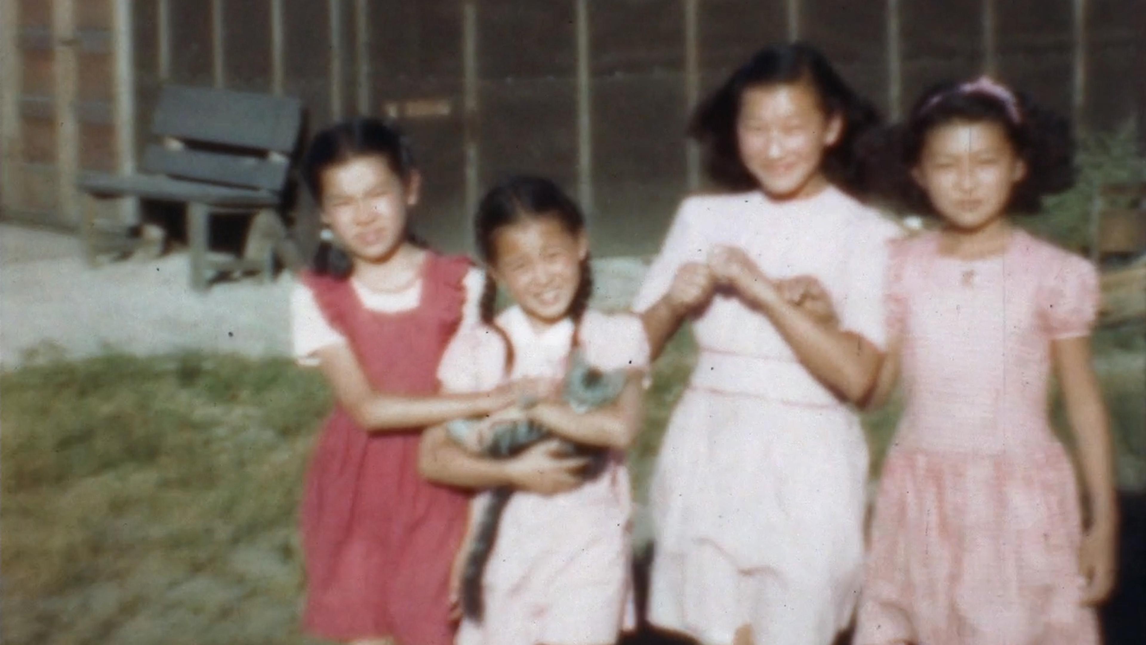 Four young girls in dresses smile outside, with one holding a small cat.