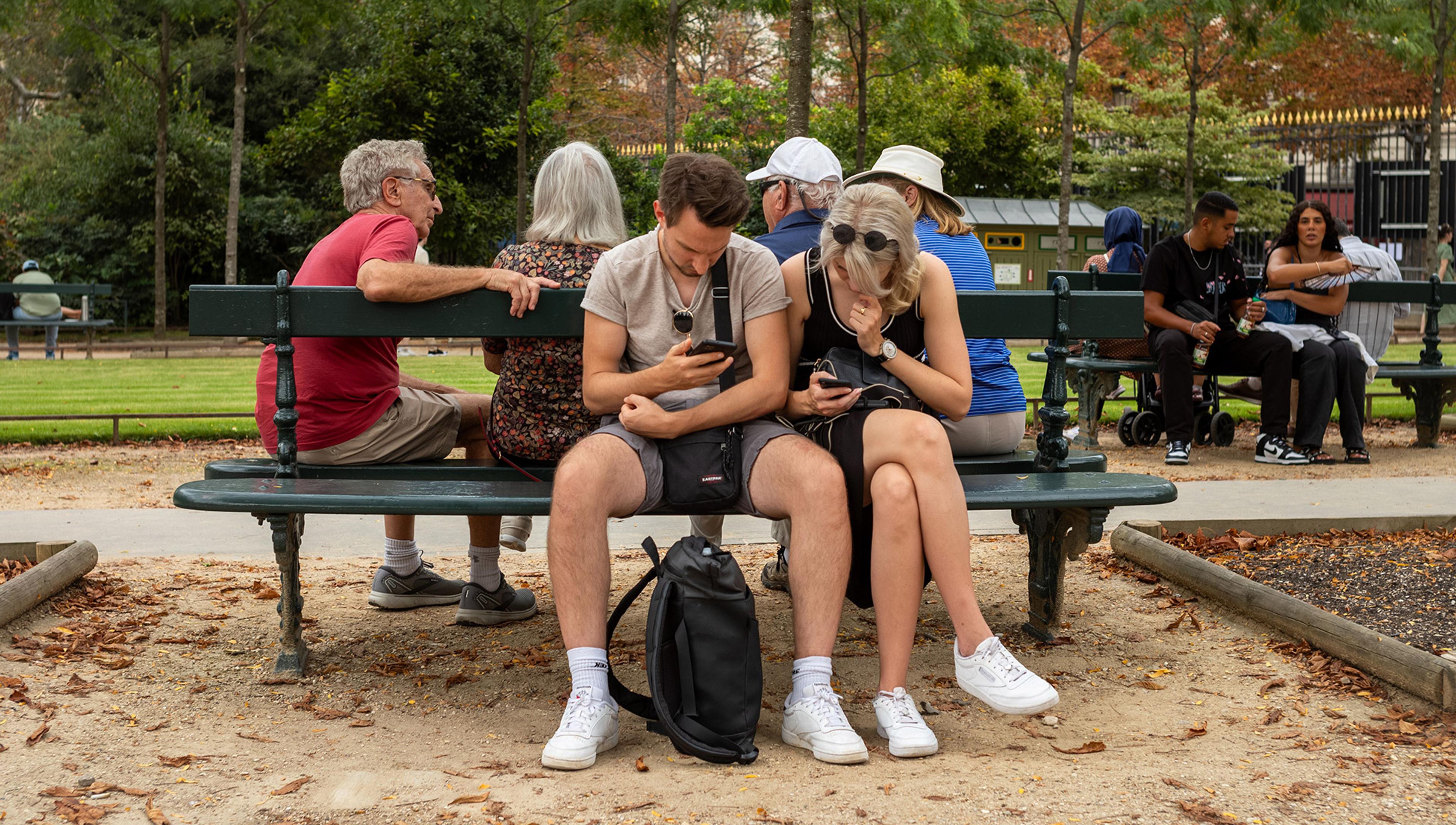 People of different ages and genders sitting on park benches, some chatting, some looking at their smartphones