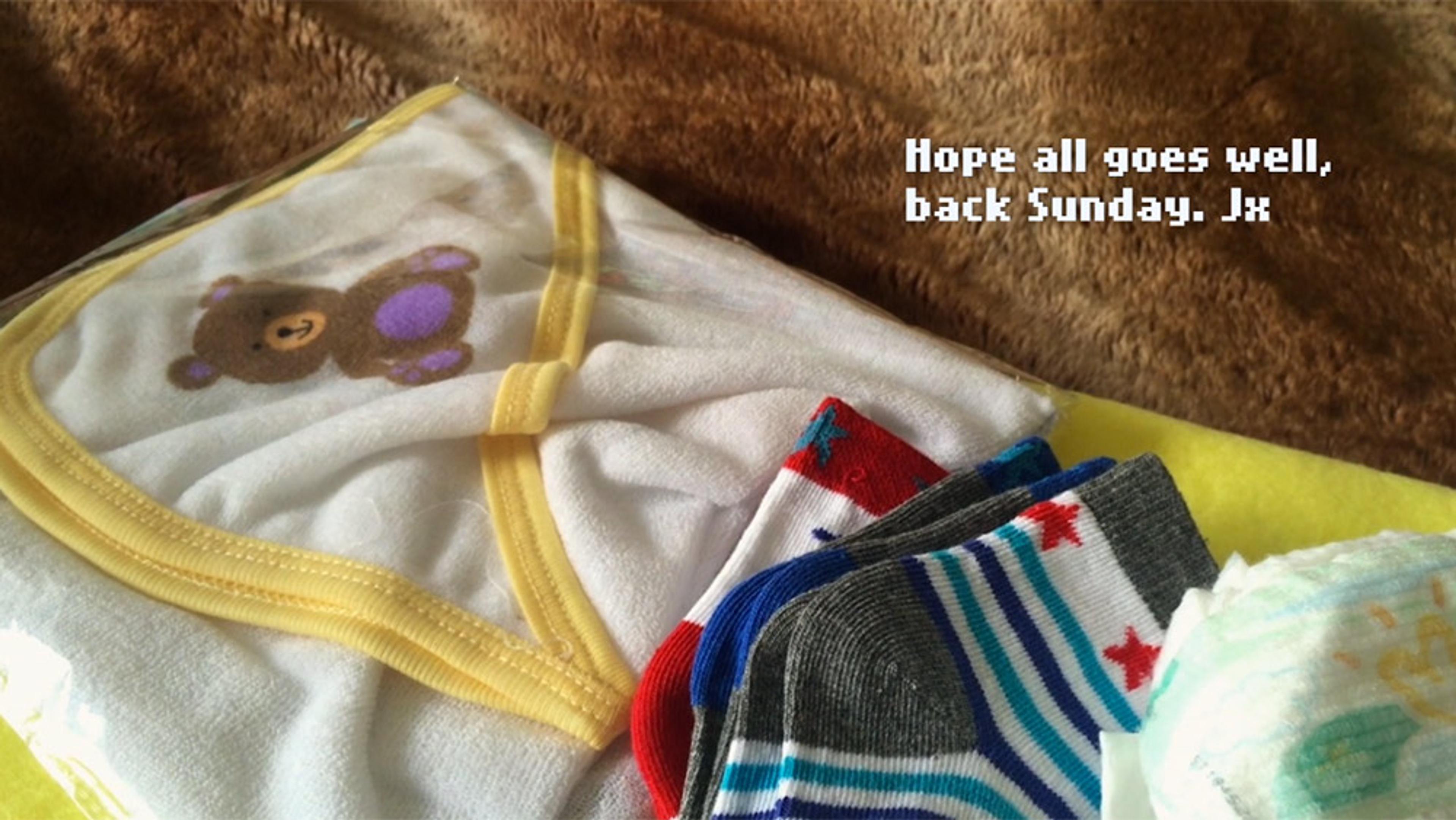 Folded baby clothes including a blanket with a bear, colourful socks, and a diaper, with text “Hope all goes well, back Sunday. Jx”.