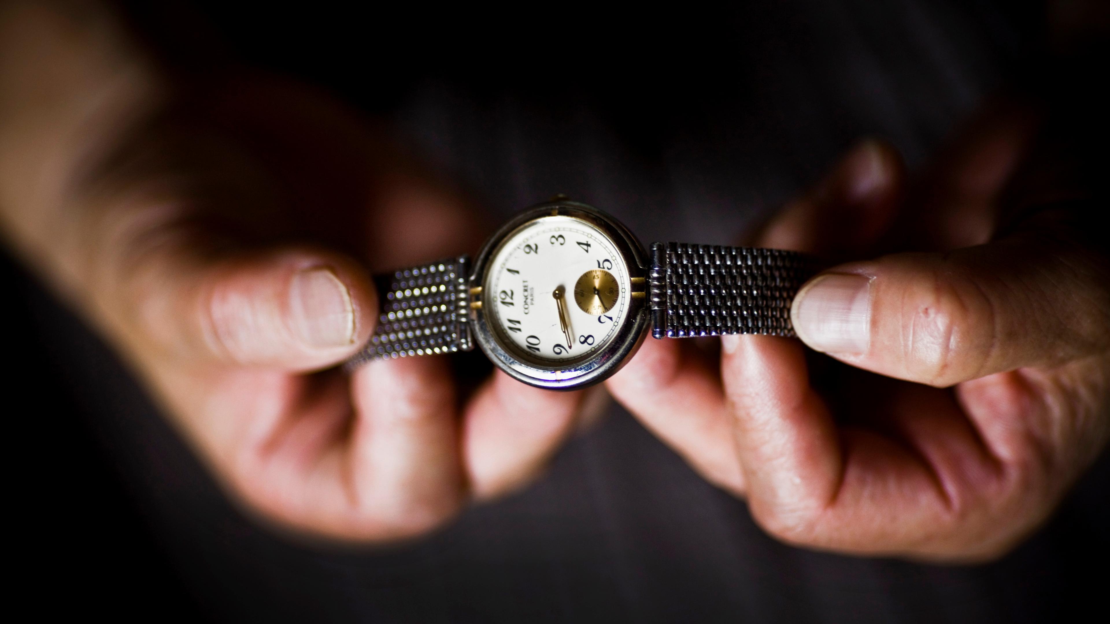 Close-up photo of hands holding a silver wristwatch with a white dial and black numerals on a black background.