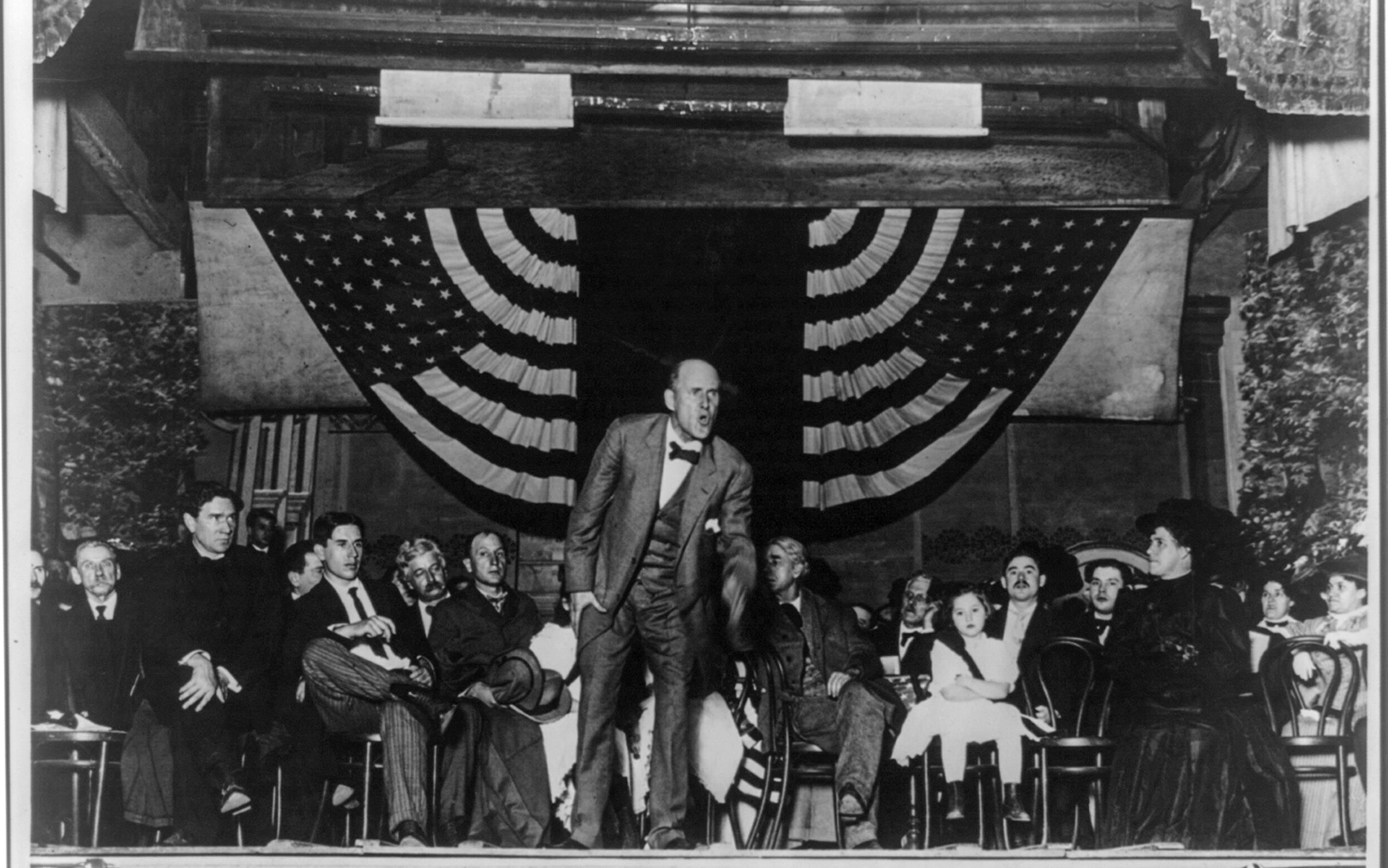 Eugene V Debs making a speech from a stage on which several other men, women and a young girl are seated behind him; also shows 48-star flags of the United States hanging at the back of the stage