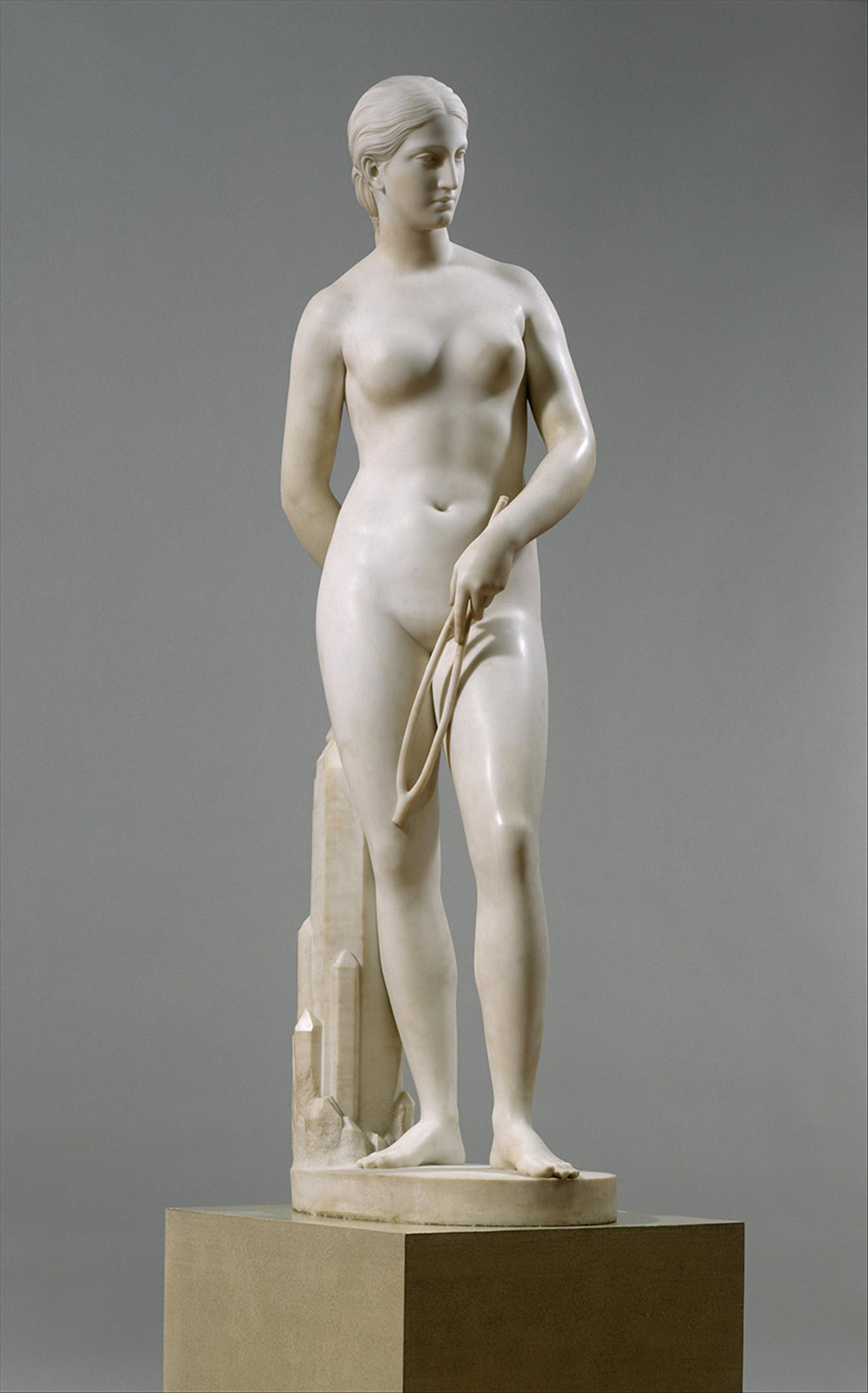 Marble statue of a nude woman holding a divining rod, standing on a pedestal against a plain grey background.