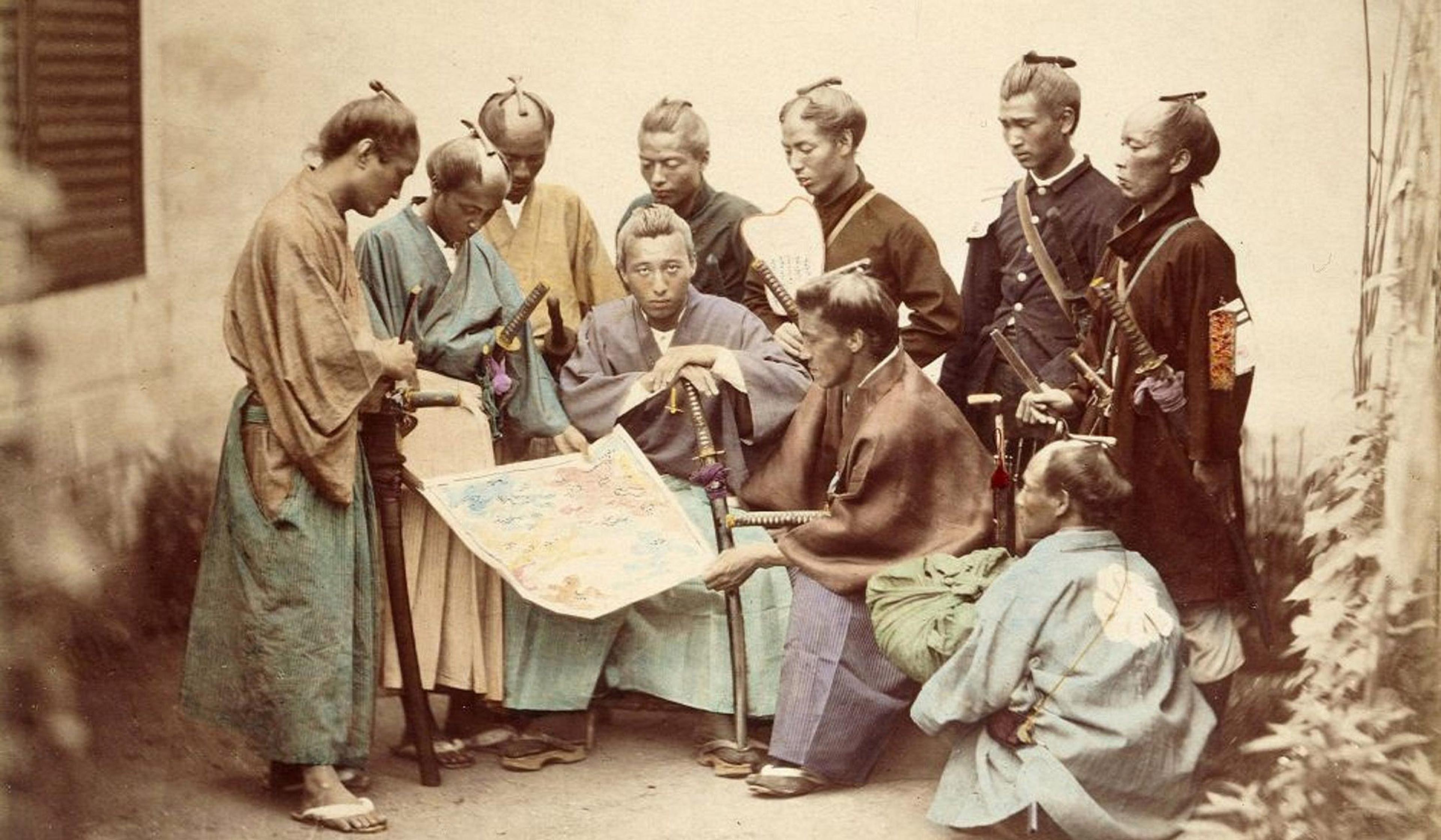 Sepia photo of a group of Japanese samurai studying a map, some standing, some sitting or kneeling, in traditional attire and swords.
