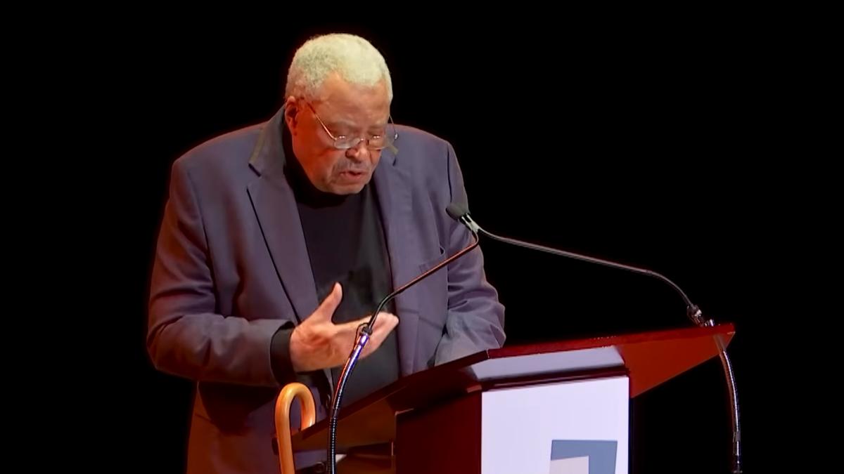 Photo of an elderly man speaking at a podium with a microphone onstage, wearing a dark suit and glasses.