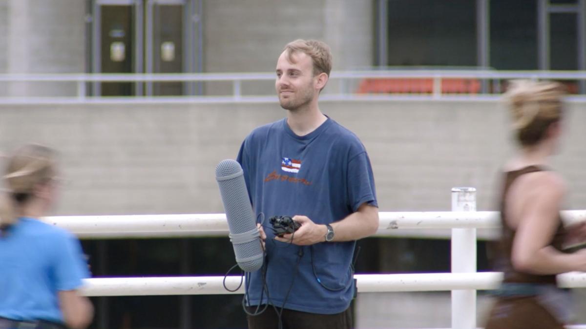 Photo of a man holding a microphone and equipment standing on a walkway with blurred people passing by in the foreground.