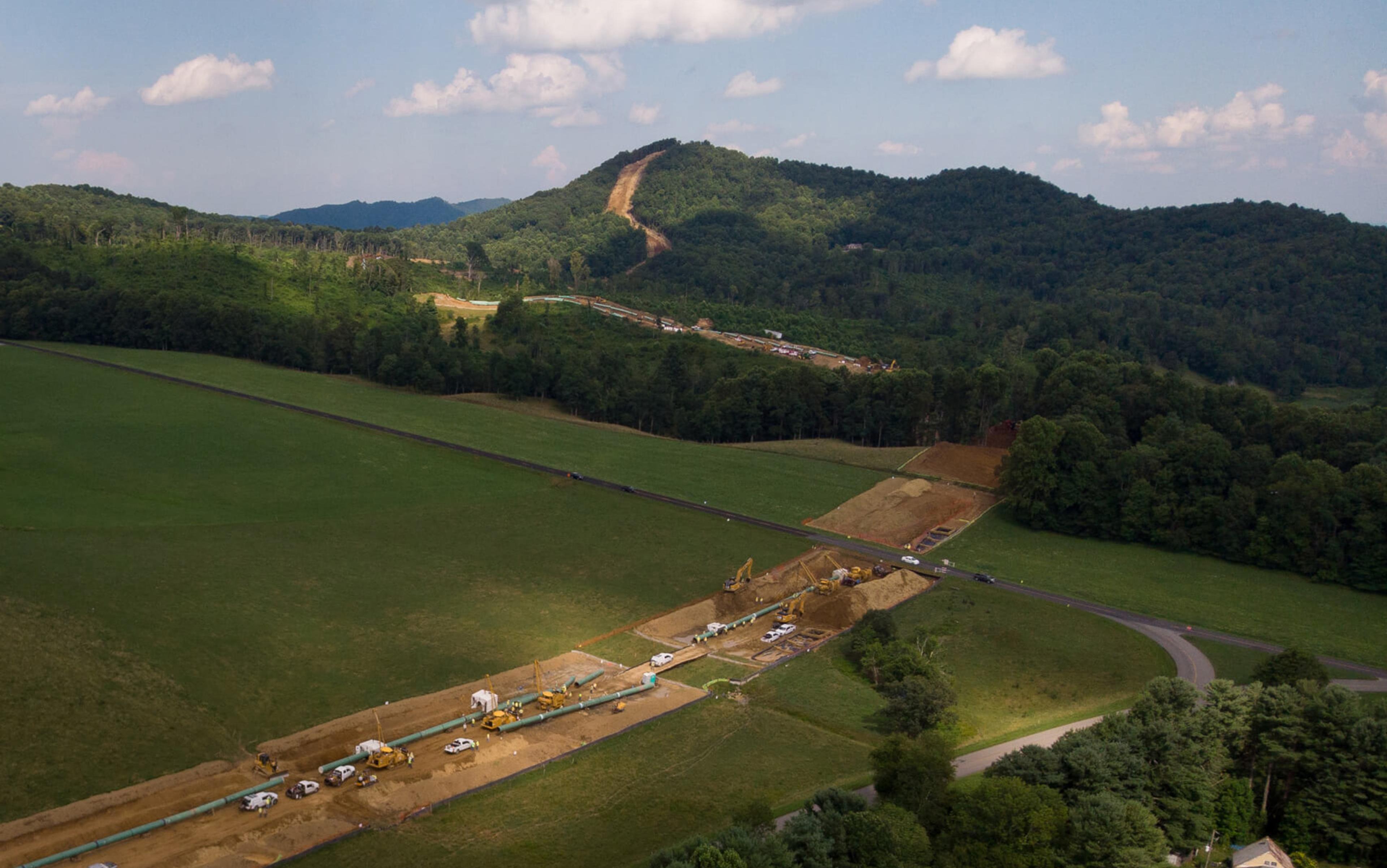 Aerial view of a large pipeline construction site with machinery and vehicles cutting through green fields and hills under a partly cloudy sky.