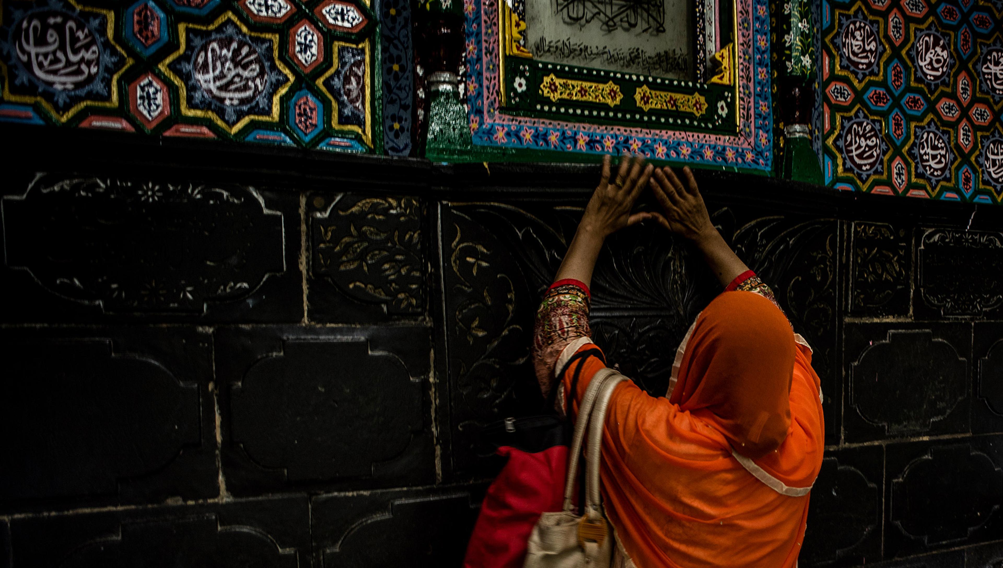 A muslim woman in a colourful orange sari reaches up to touch a decorative panel above a shrine