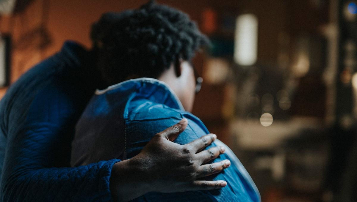 Photo of two people embracing indoors, one hand resting gently on the other’s back, both wearing blue clothing with soft-focus background.