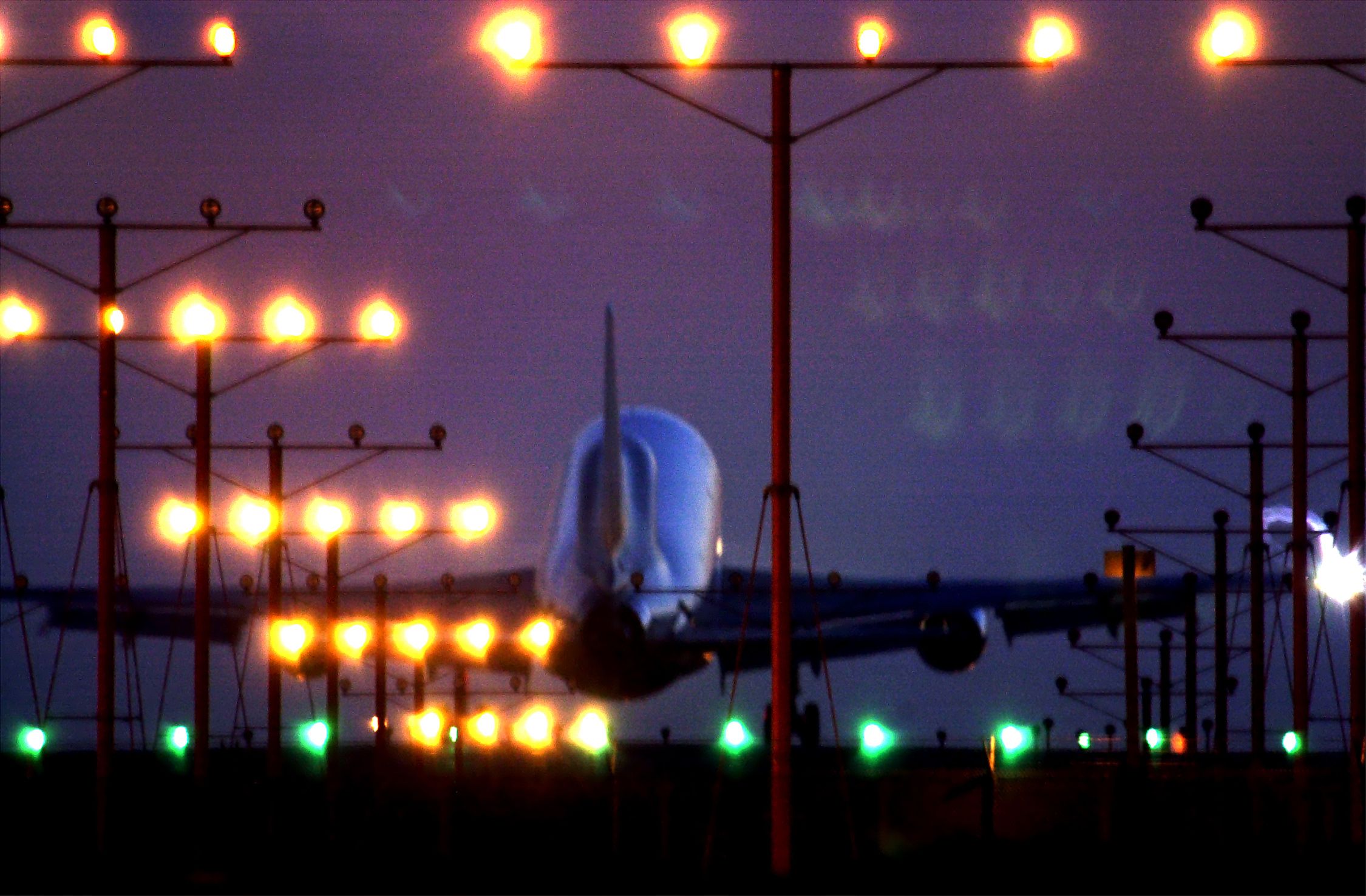 An aeroplane landing at dusk with runway lights glowing, surrounded by a purple sky and silhouetted light poles.