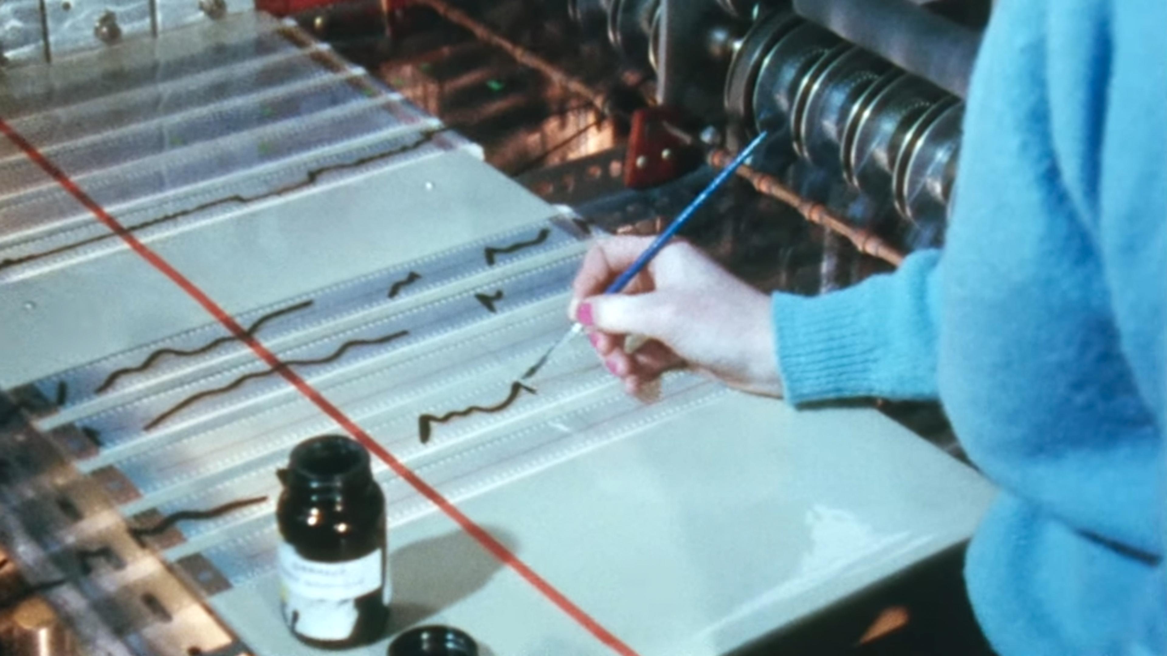 Photo of a person painting waveforms on clear film with a brush and ink beside audio equipment.