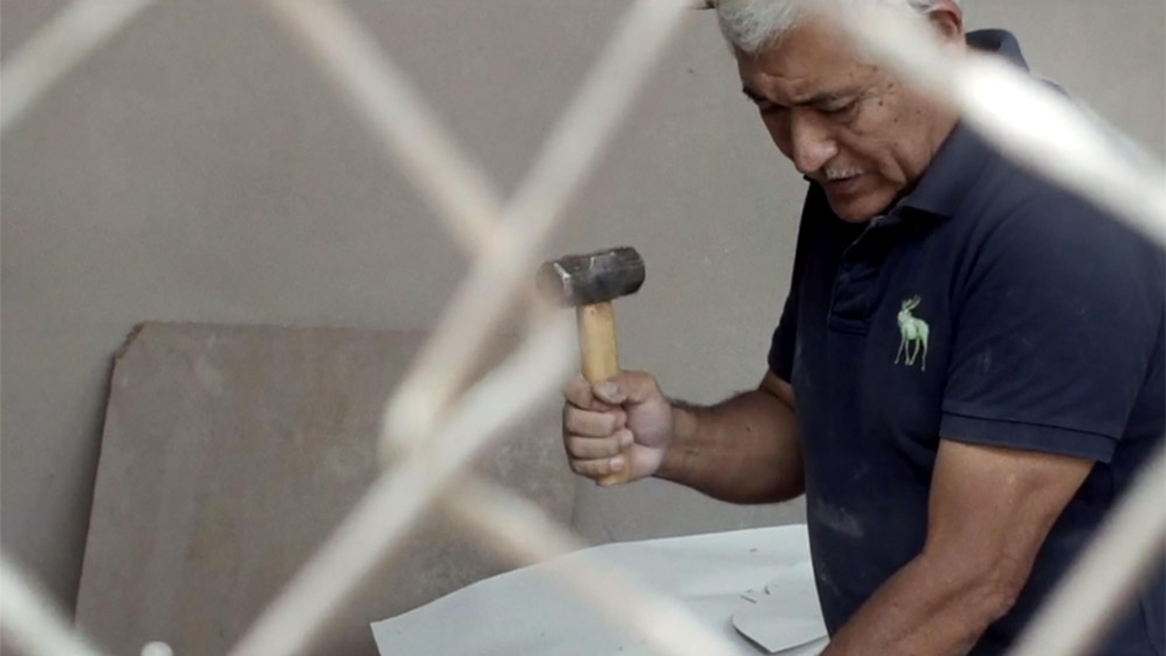 A man in a dark blue shirt hammering an object with a mallet, viewed through a chain-link fence.