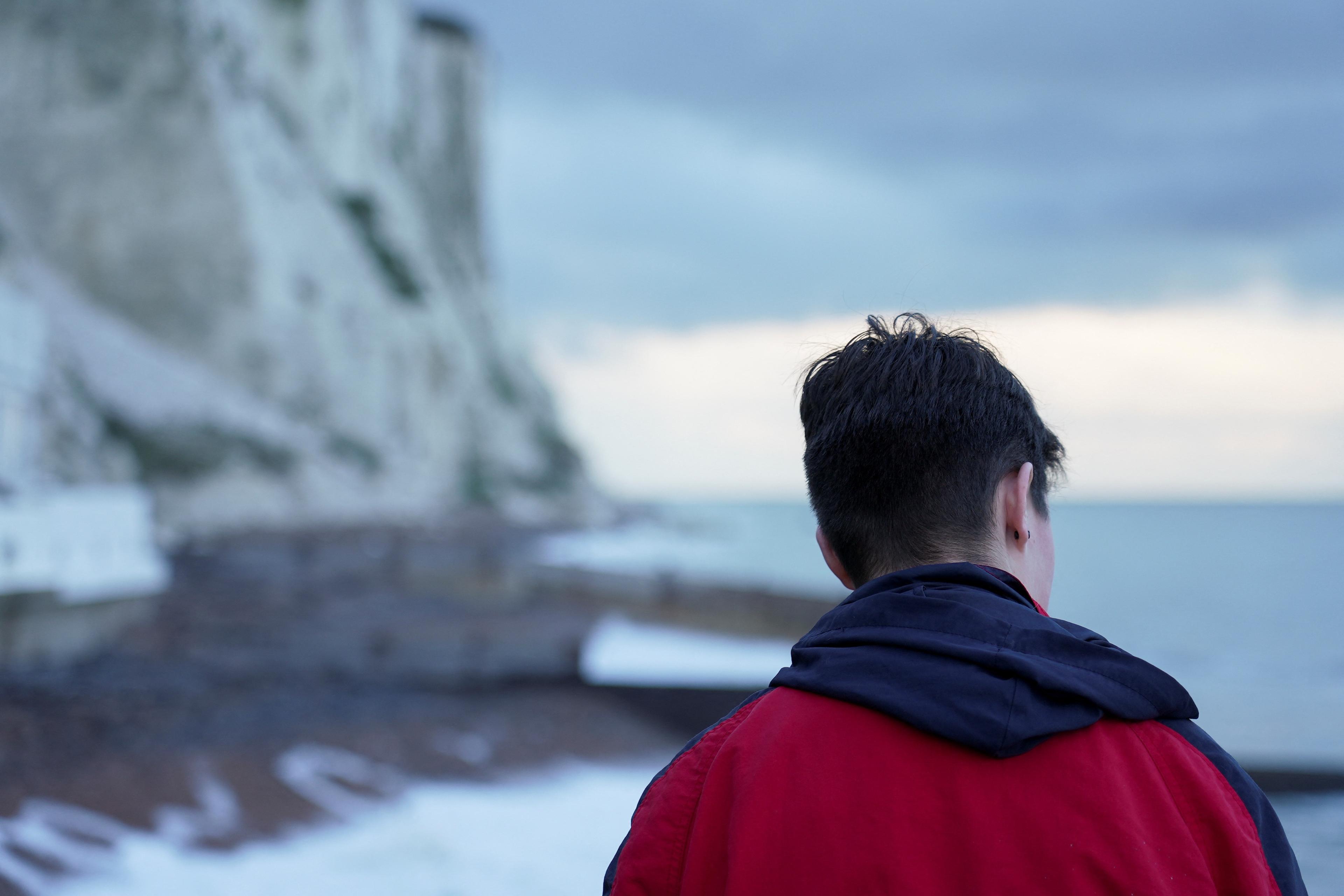 Photo of a person in a red jacket looking at cliffs by the sea under a cloudy sky.