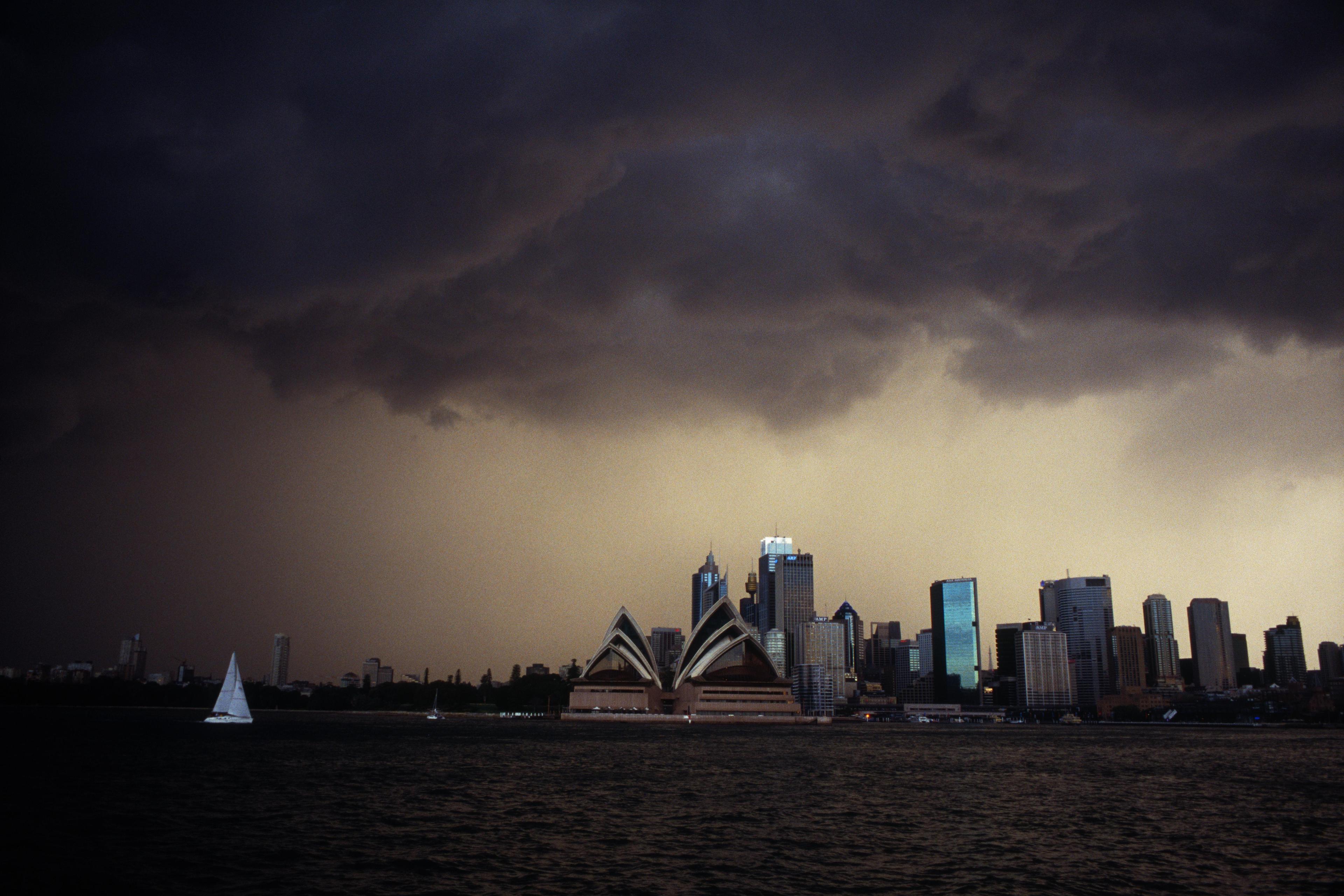 Photo of a stormy sky over Sydney Opera House and city skyline with dark clouds and a sailboat on the water.