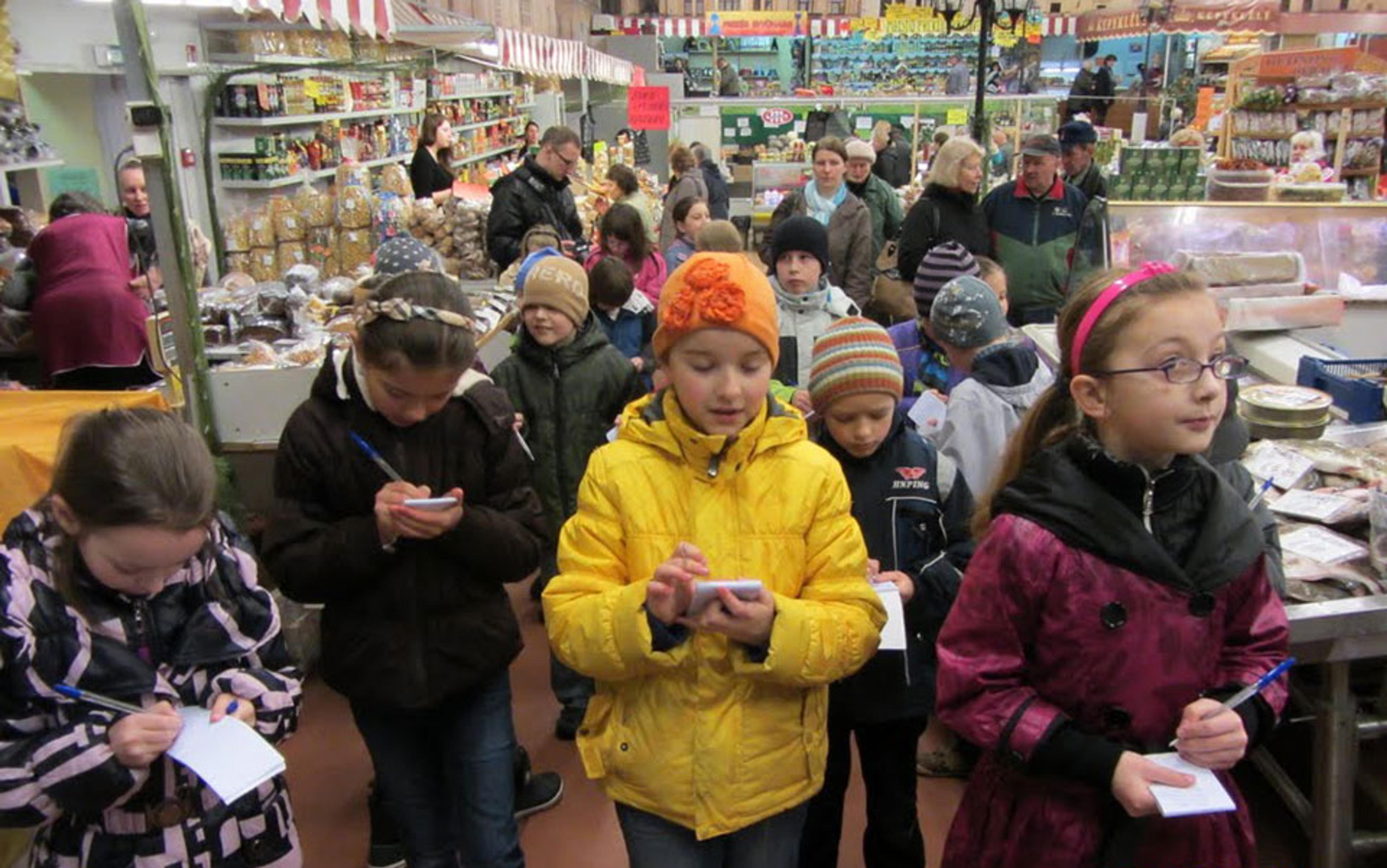 Children in winter clothing writing on notepads in a crowded indoor market with food stalls in the background.